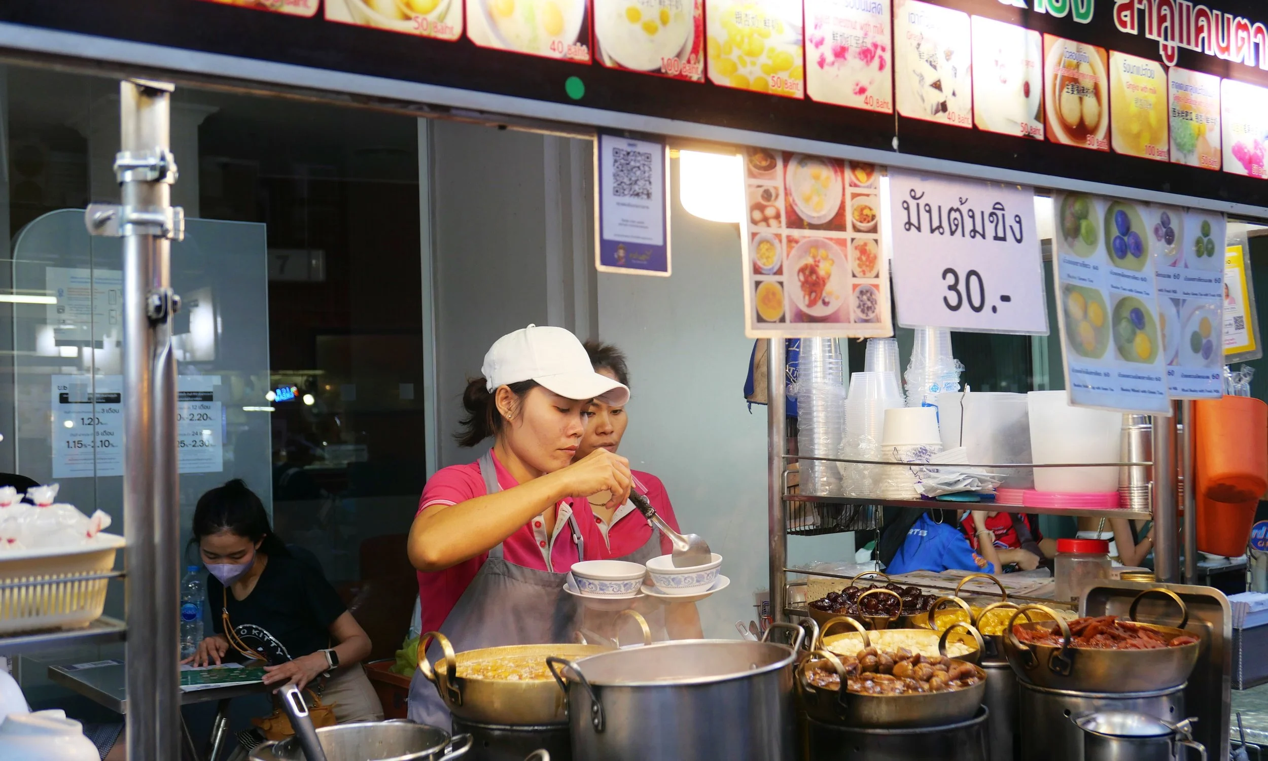 A street food vendor preparing bowls of food at an outdoor stall. The vendor is wearing a pink shirt and a white cap, with various dishes and ingredients displayed around. There are signs and menu boards in the background with prices in Thai Baht.