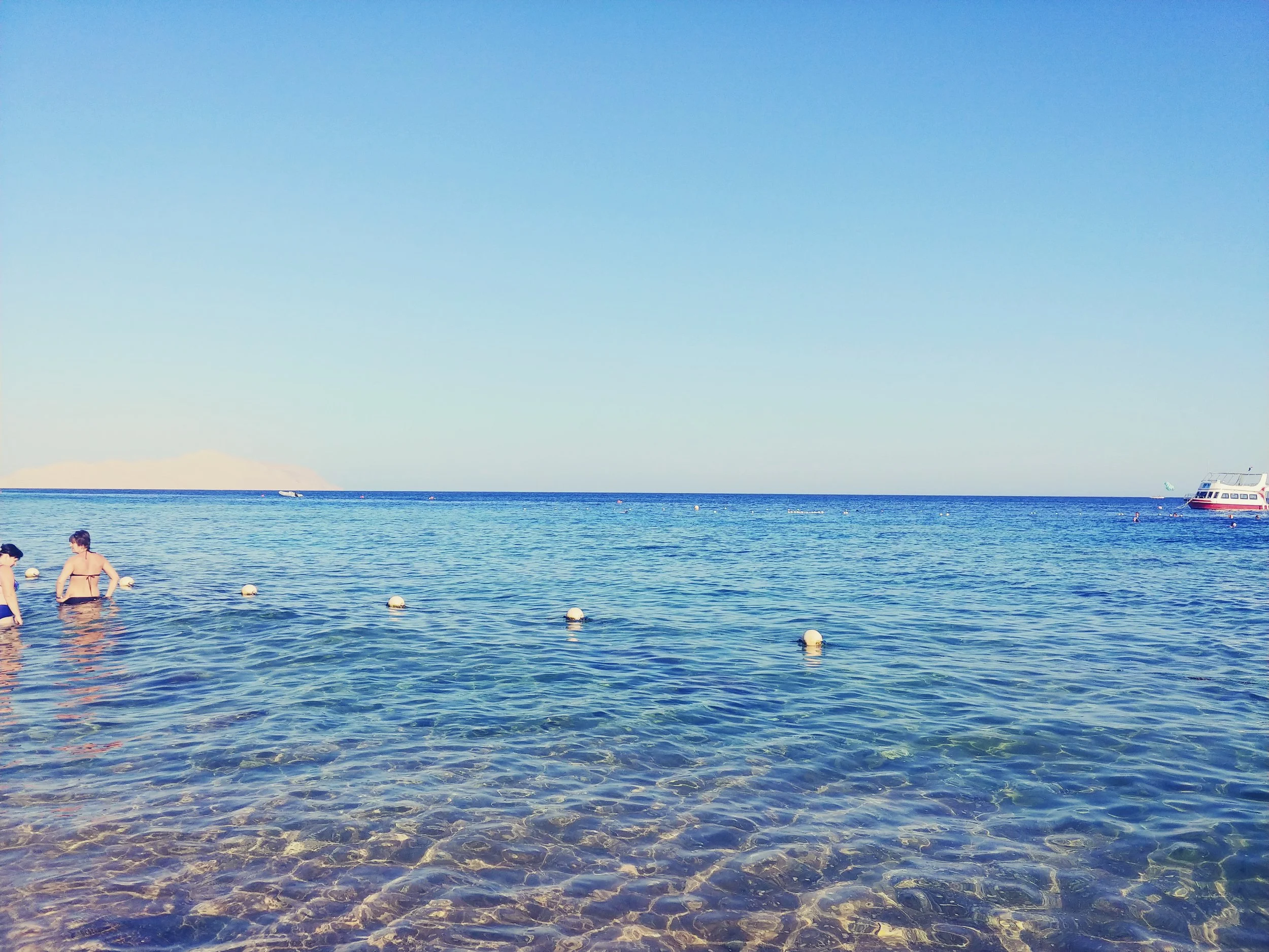 People swimming and wading in the clear, blue ocean with a boat docked nearby, under a bright, clear sky.