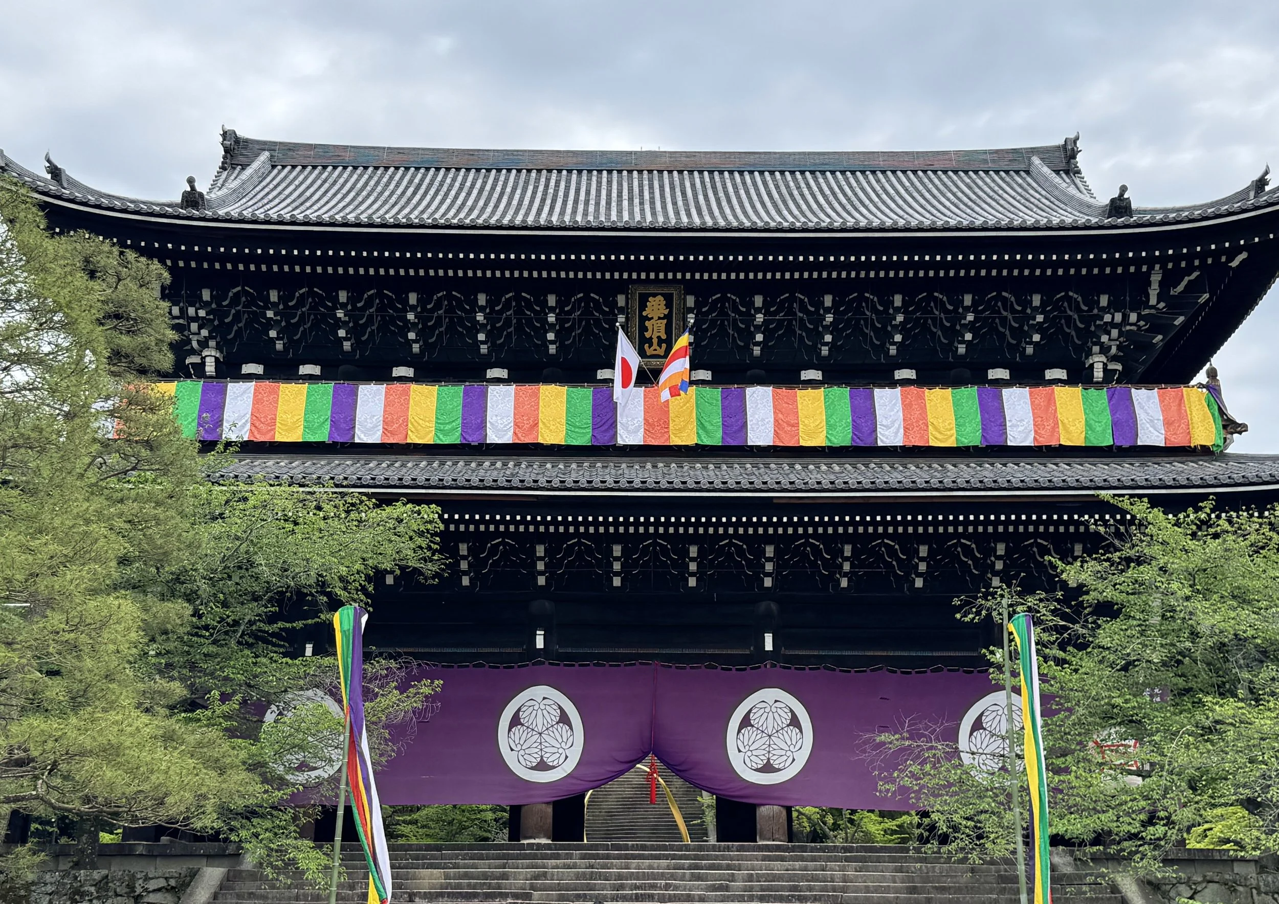 A traditional Japanese temple with a arched tiled roof, decorated with colorful festival banners in green, purple, white, yellow, and red. A purple curtain with white circular patterns hangs at the entrance, and there are steps leading up to the temp