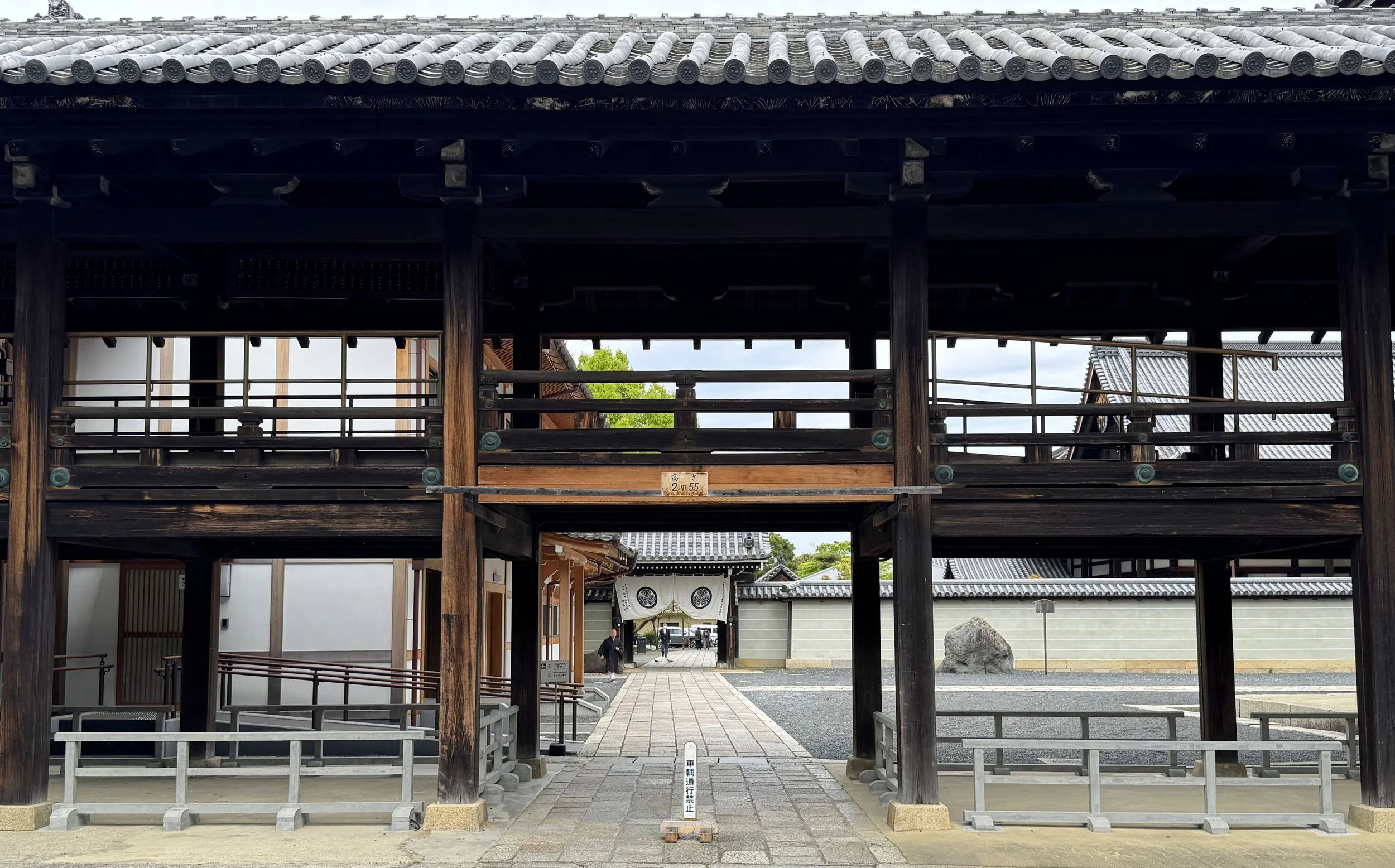 Traditional Japanese temple entrance with wooden gate, stone pathway, and tiled roofs, with a few people walking in the background.