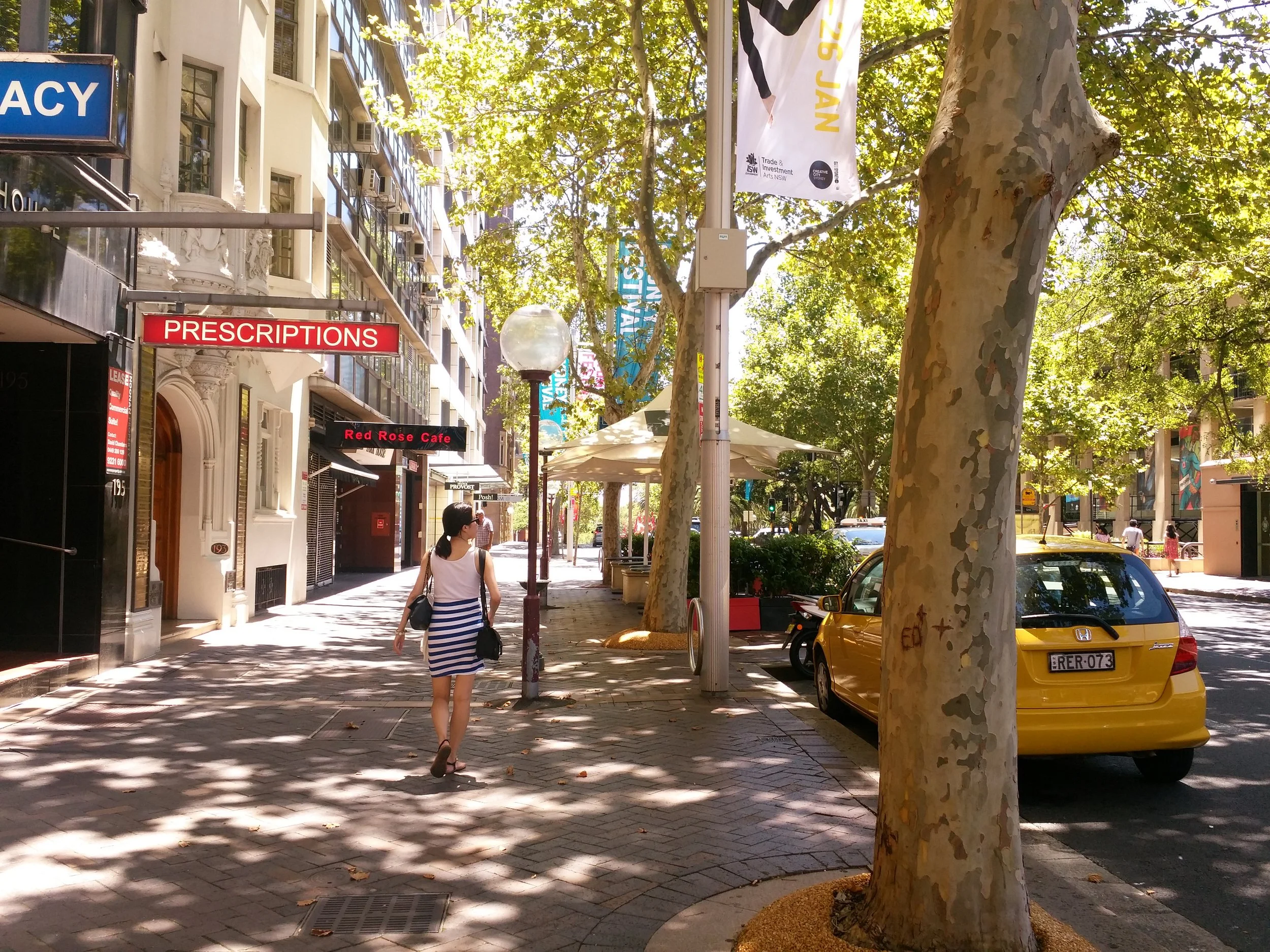 A city sidewalk with trees, a woman in a blue and white striped dress walking, and parked yellow cars. Buildings and signs, including one that says 'PRESCRIPTIONS,' are visible in the background.