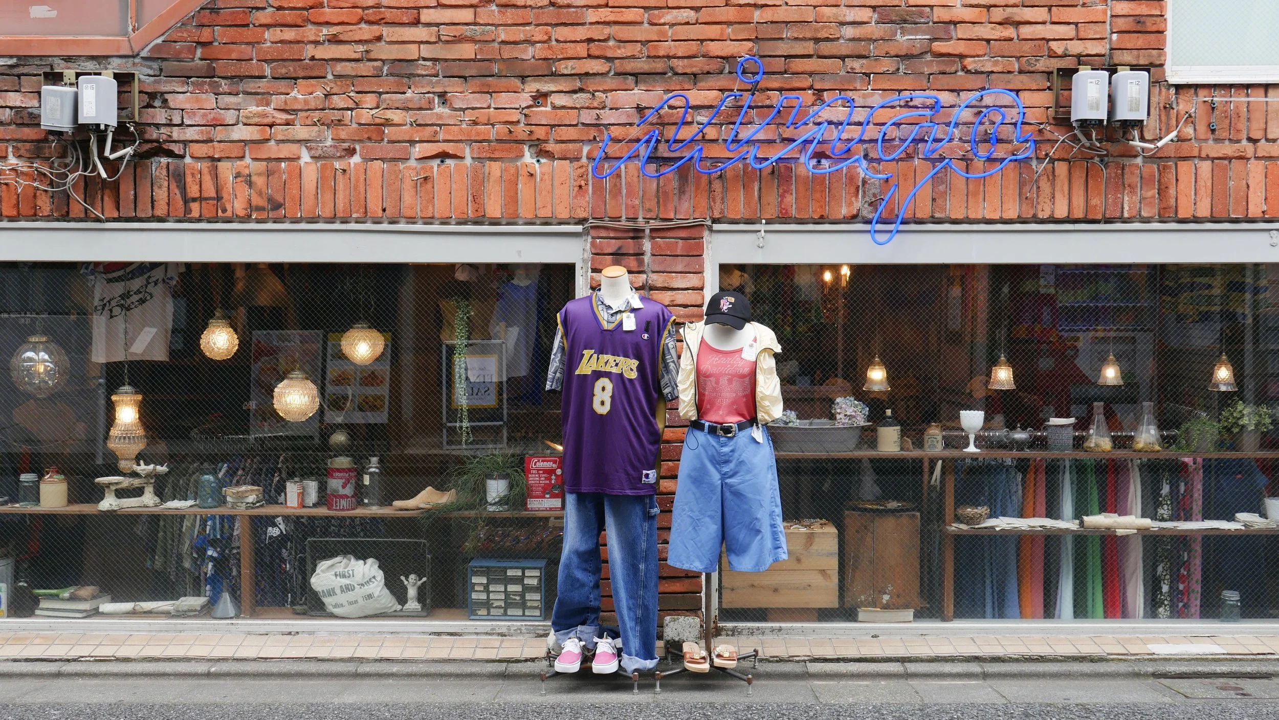 Storefront with two mannequins dressed in casual clothes, one wearing a purple Lakers jersey and denim jeans, the other in a white jacket and pink shirt. The store has a brick exterior with various items displayed inside, including lamps and plants. 