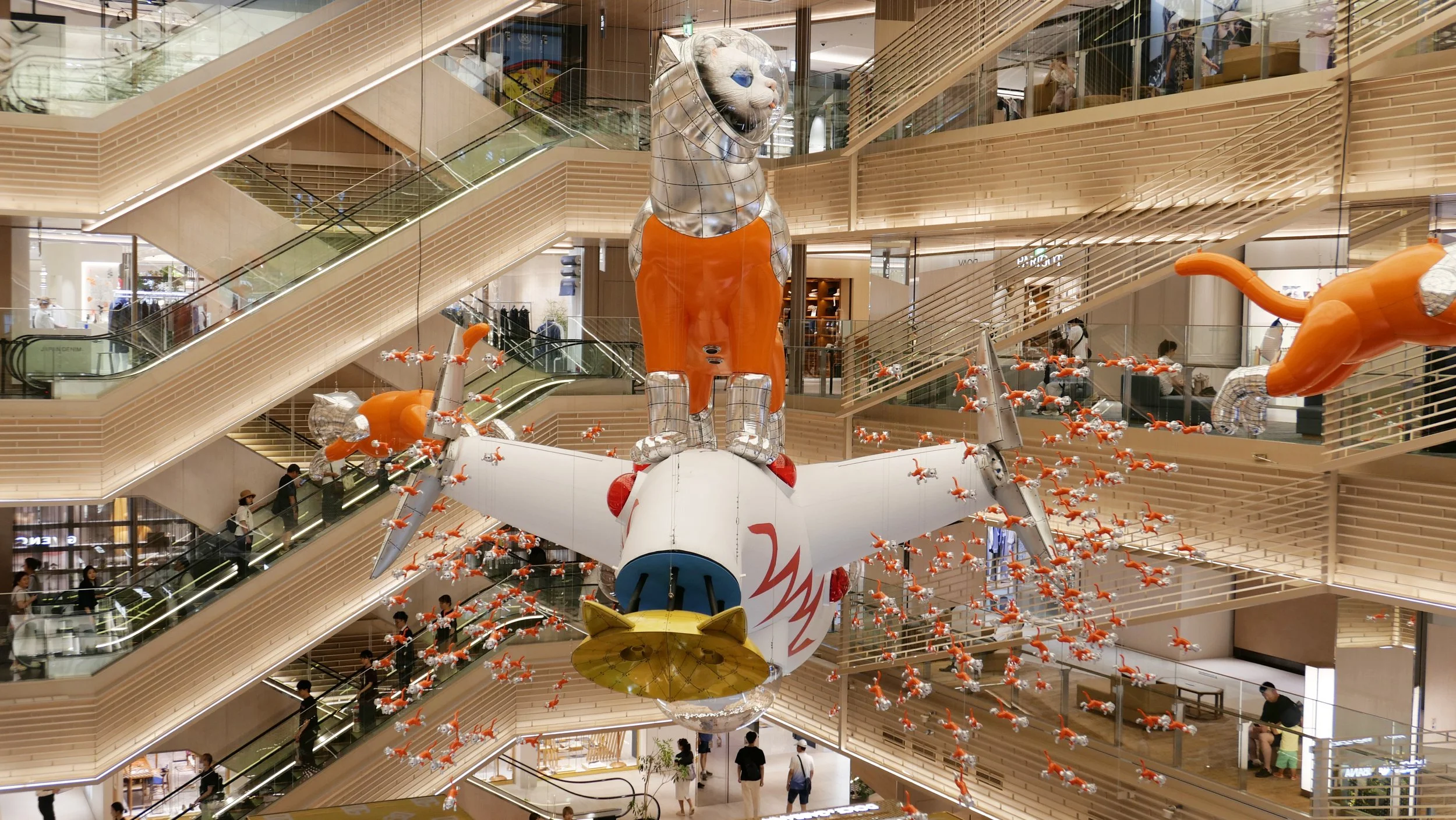 Large decorative sculpture of a mouse with silver head and orange body, surrounded by numerous small orange mice suspended in midair inside a multi-floor shopping mall with escalators and shoppers.