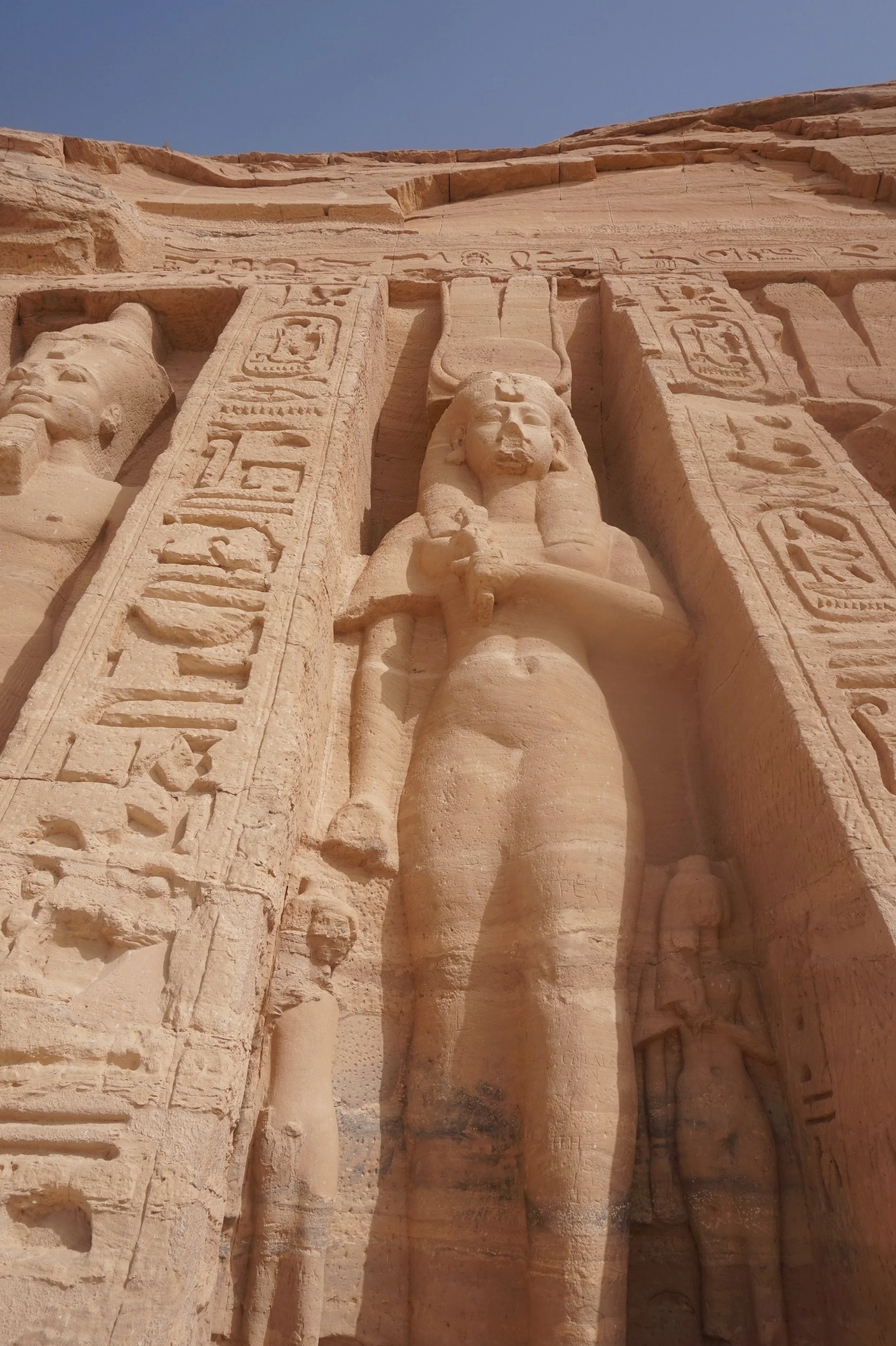 Close-up of ancient Egyptian stone carvings on a large monument, including depictions of figures, hieroglyphics, and a face with a headdress, against a clear blue sky.