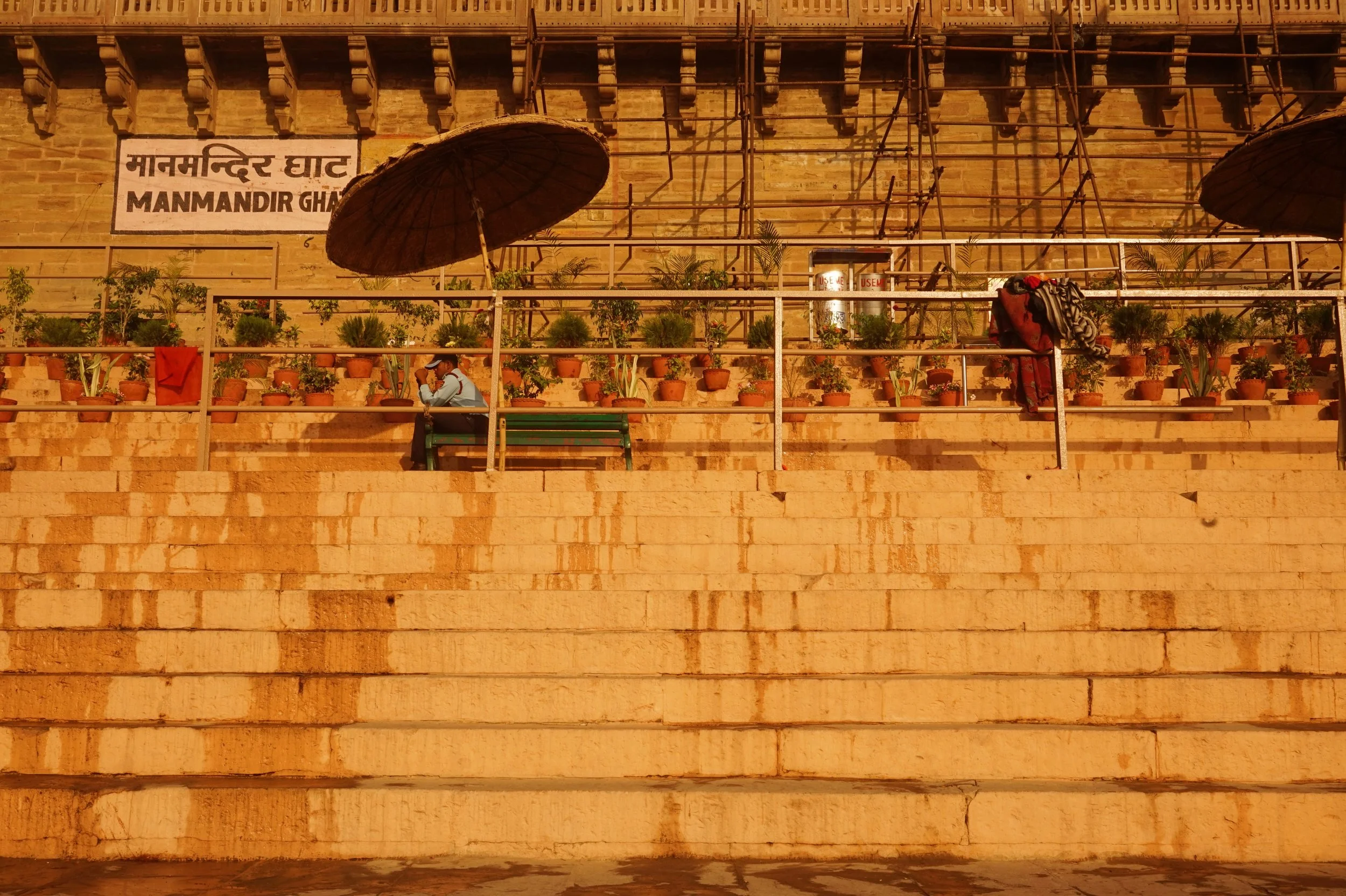 The image shows the steps of the Manmandir Ghat with pots of plants arranged on the railings and two large umbrellas providing shade, set against the historic stone wall of the ghat.