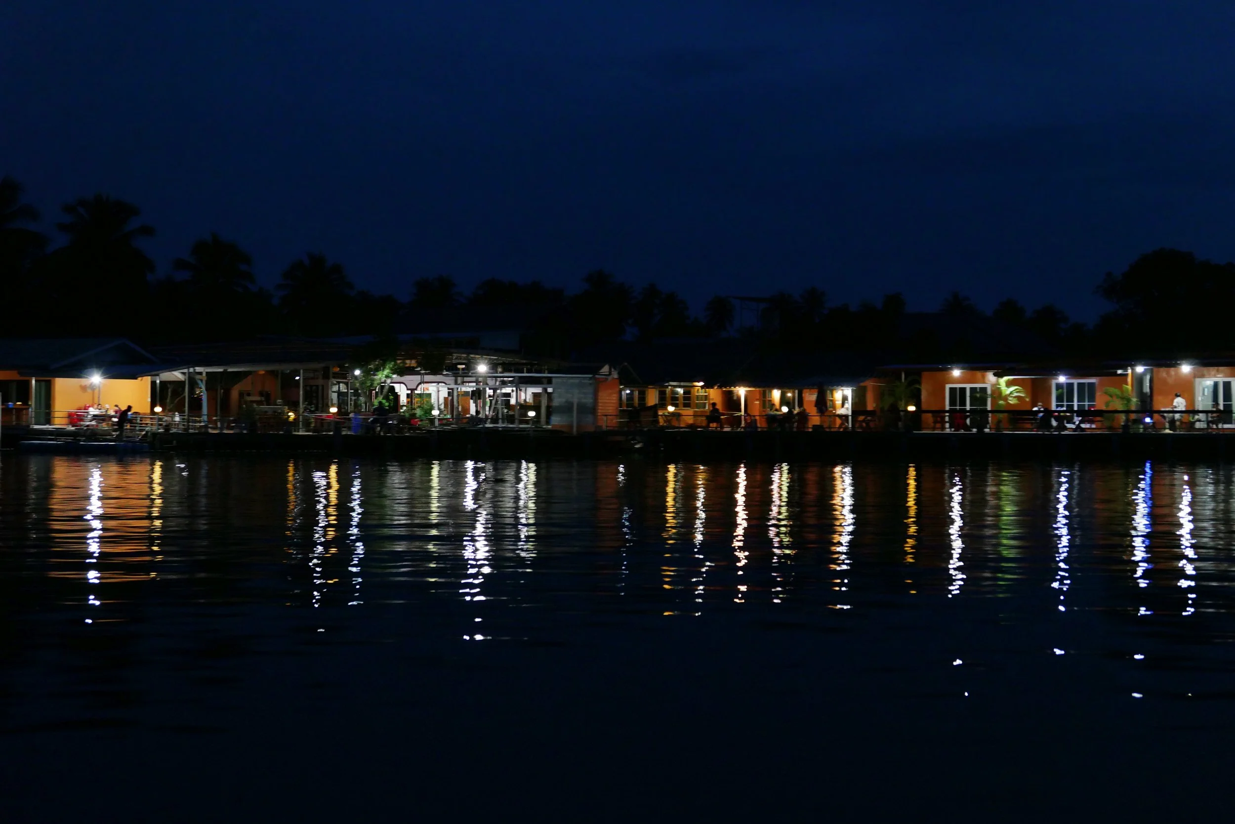 Nighttime view of brightly lit buildings and palm trees along a waterfront, with reflections shimmering on the water.