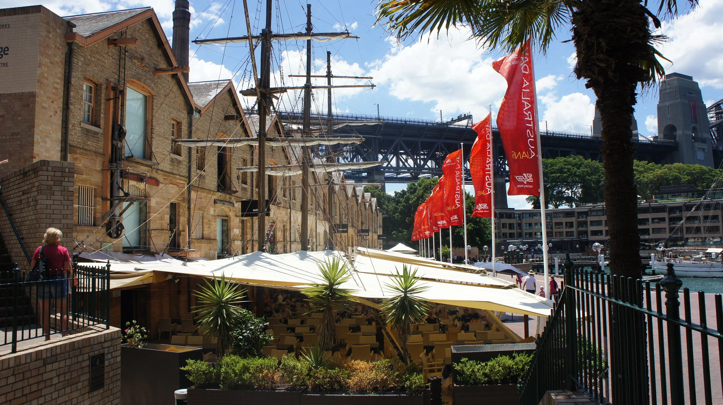 Scene of a waterfront area with historic brick buildings, flags, and a dock with boats. There is a tall ship with masts and rigging, modern bridge in the background, and people walking or sitting at outdoor tables under umbrellas.