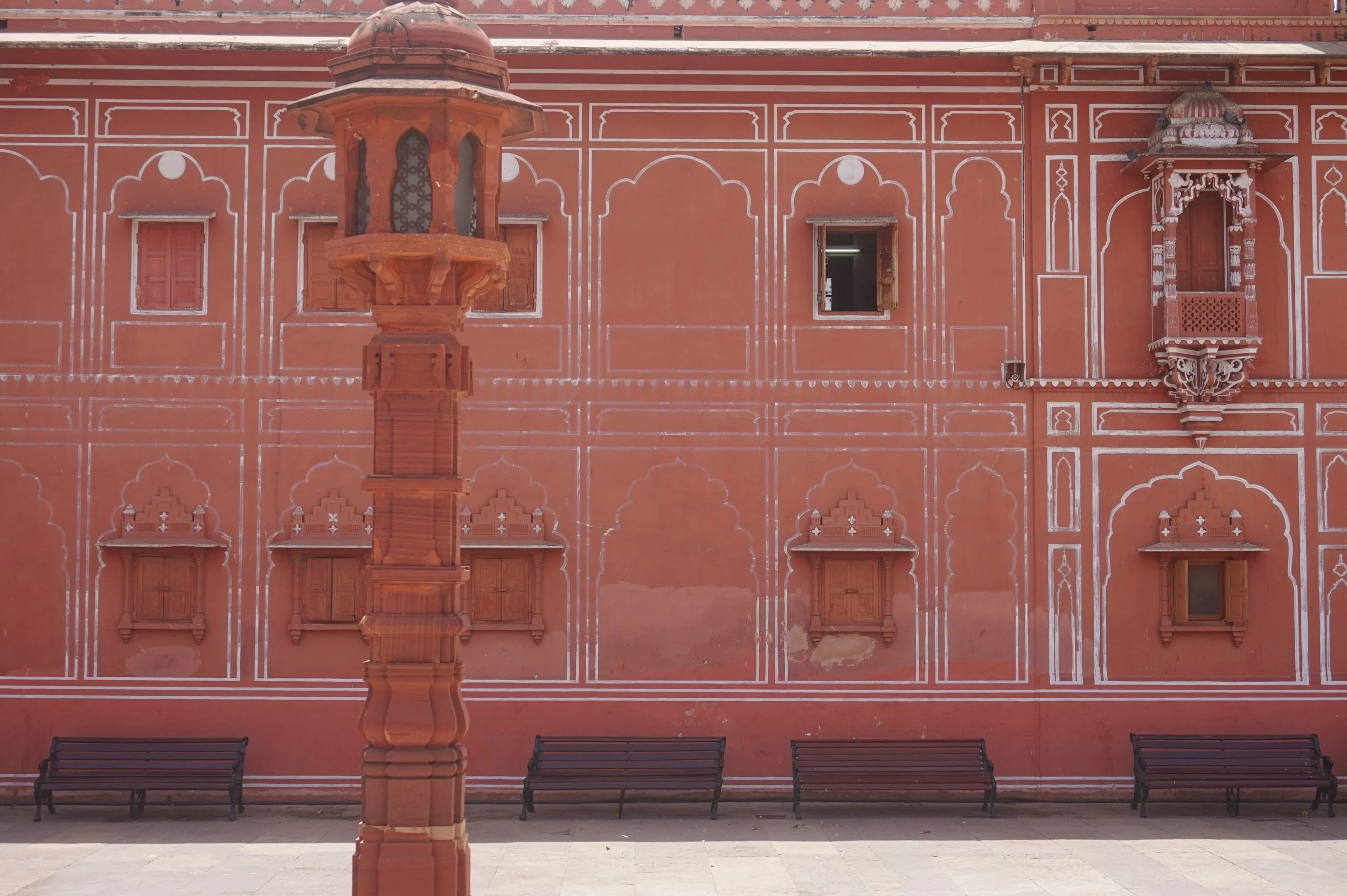A red-orange historical building with intricate white detailing and small windows, featuring two benches in front. A red stone lantern stands in the foreground.