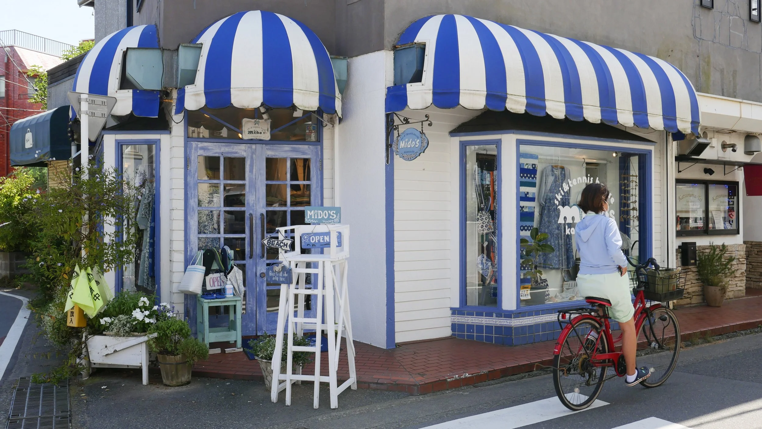 A woman wearing a light blue jacket and white shorts is riding a red bicycle on a city sidewalk in front of a shop with blue and white striped awnings. The shop's windows display various items, and there are potted plants and signs outside, including