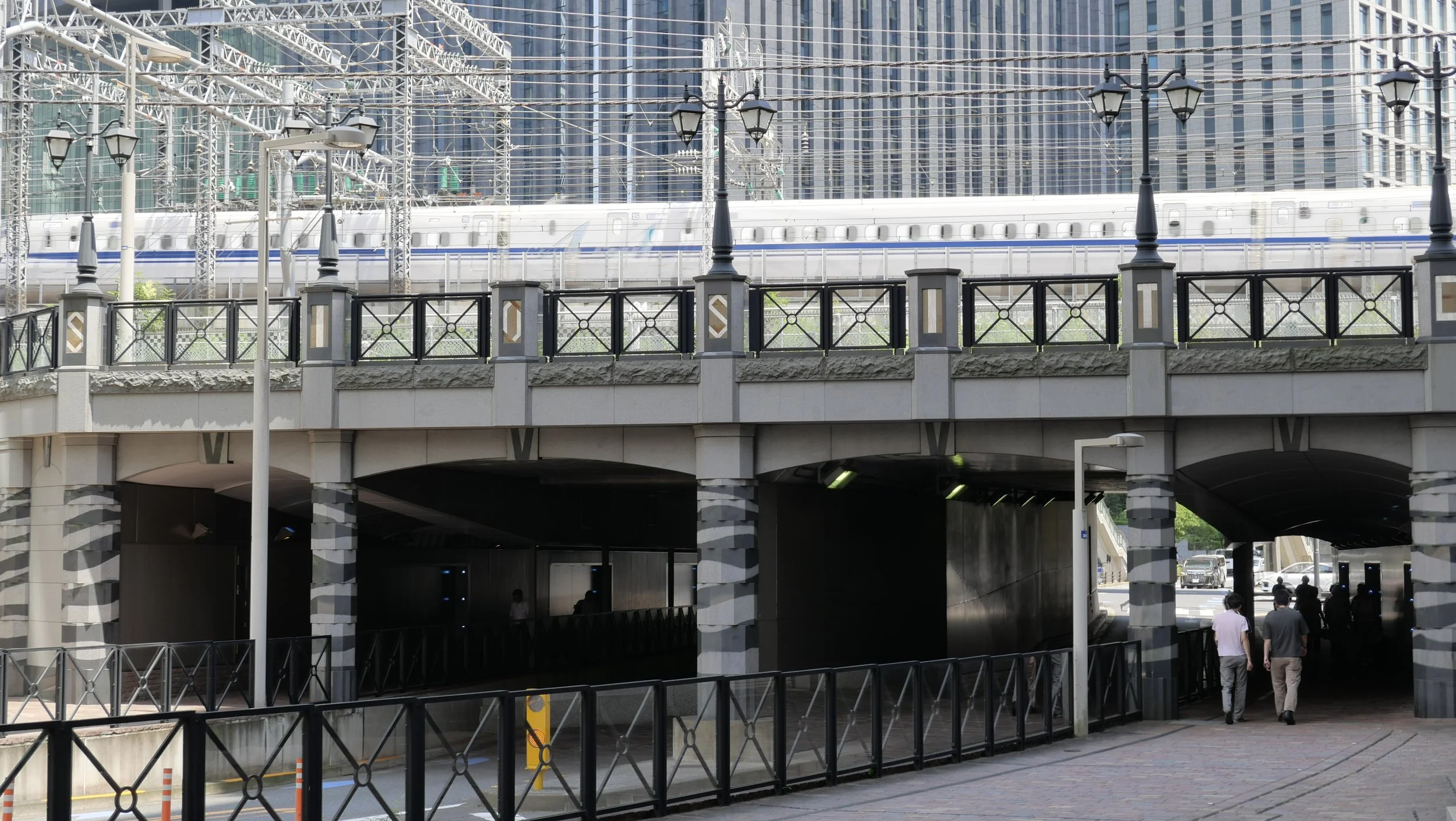 A view of an urban area with a bridge underpass, several people walking, a train on elevated tracks, and tall buildings with glass facades in the background.