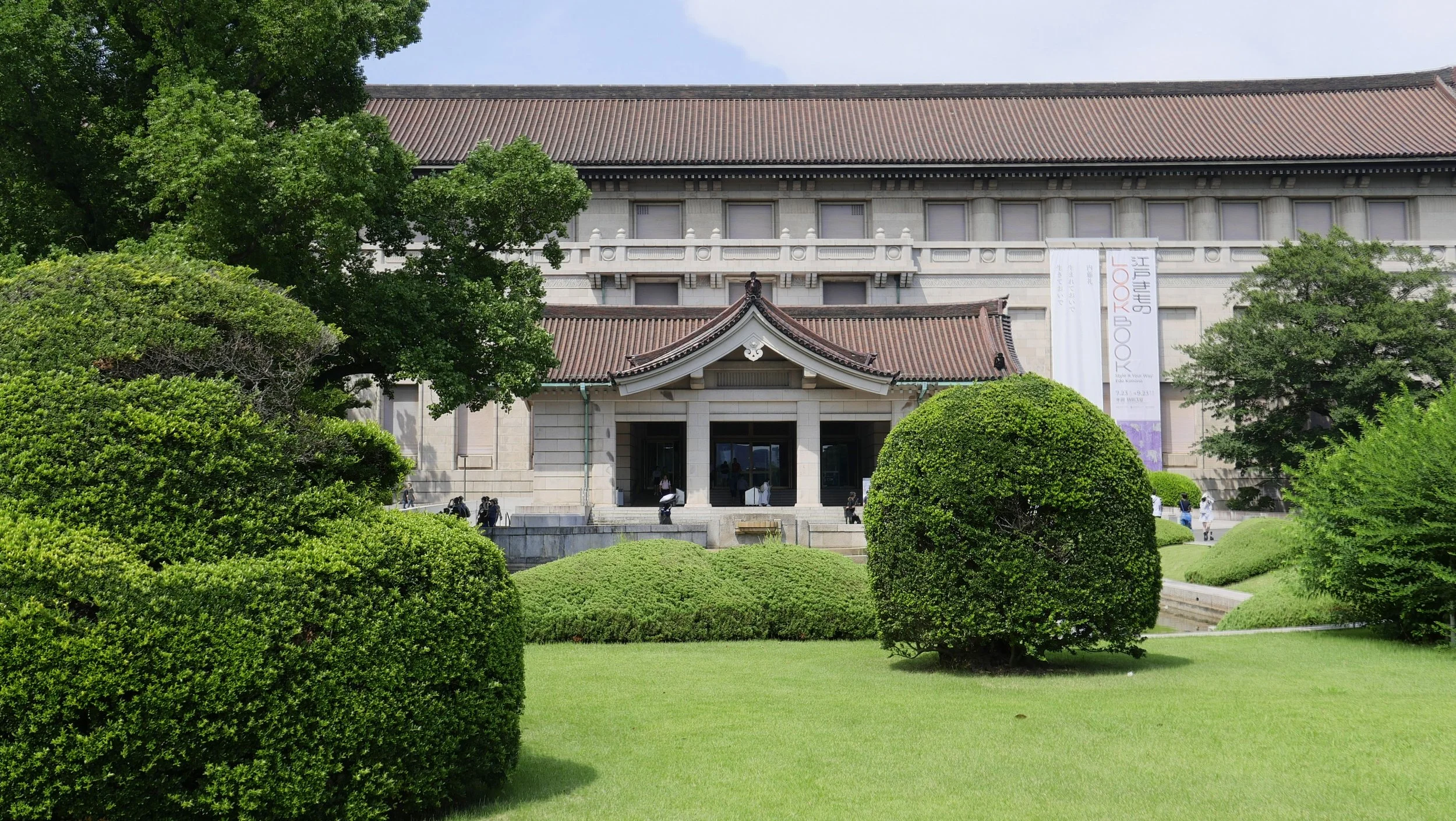 A traditional Japanese-style building with a tiled roof, surrounded by lush green bushes and trees, with a manicured lawn in the foreground. People are walking near the entrance.
