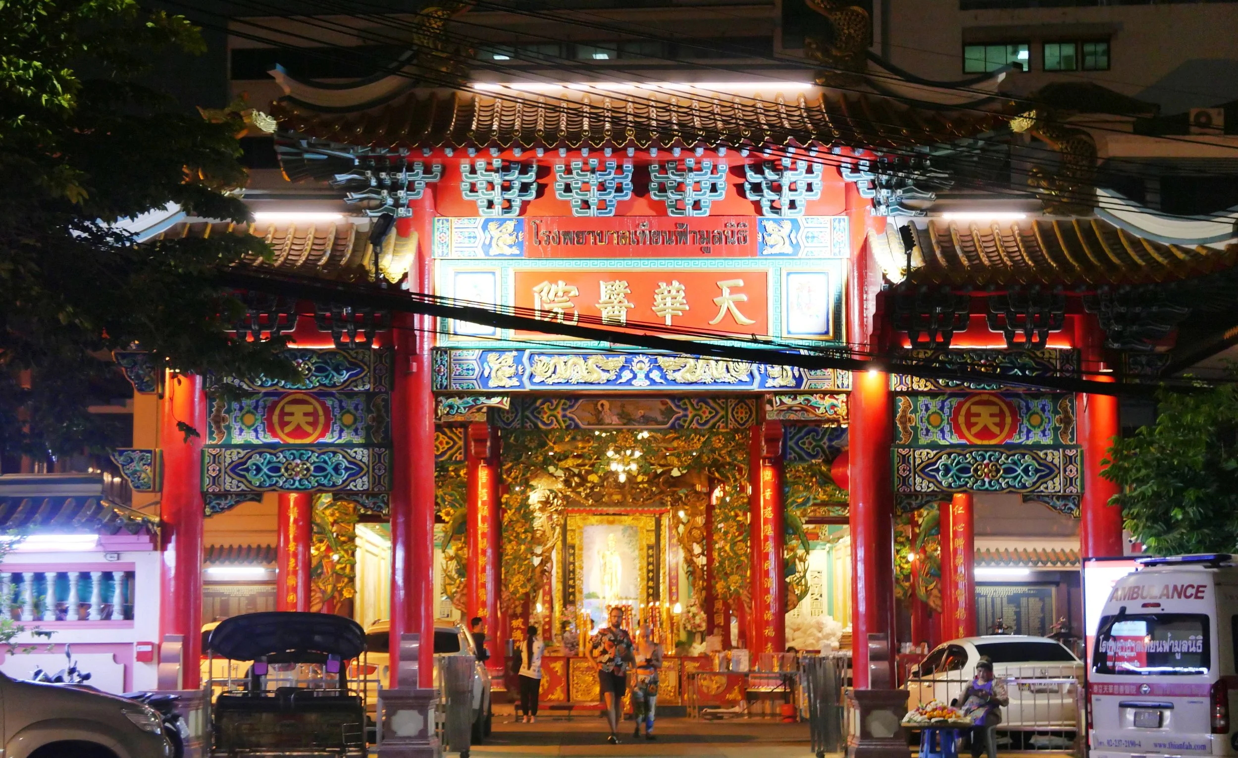 Nighttime view of a brightly lit Chinese temple entrance with red columns, ornate decorations, and golden Chinese characters. There are parked cars in front and some people near the entrance.
