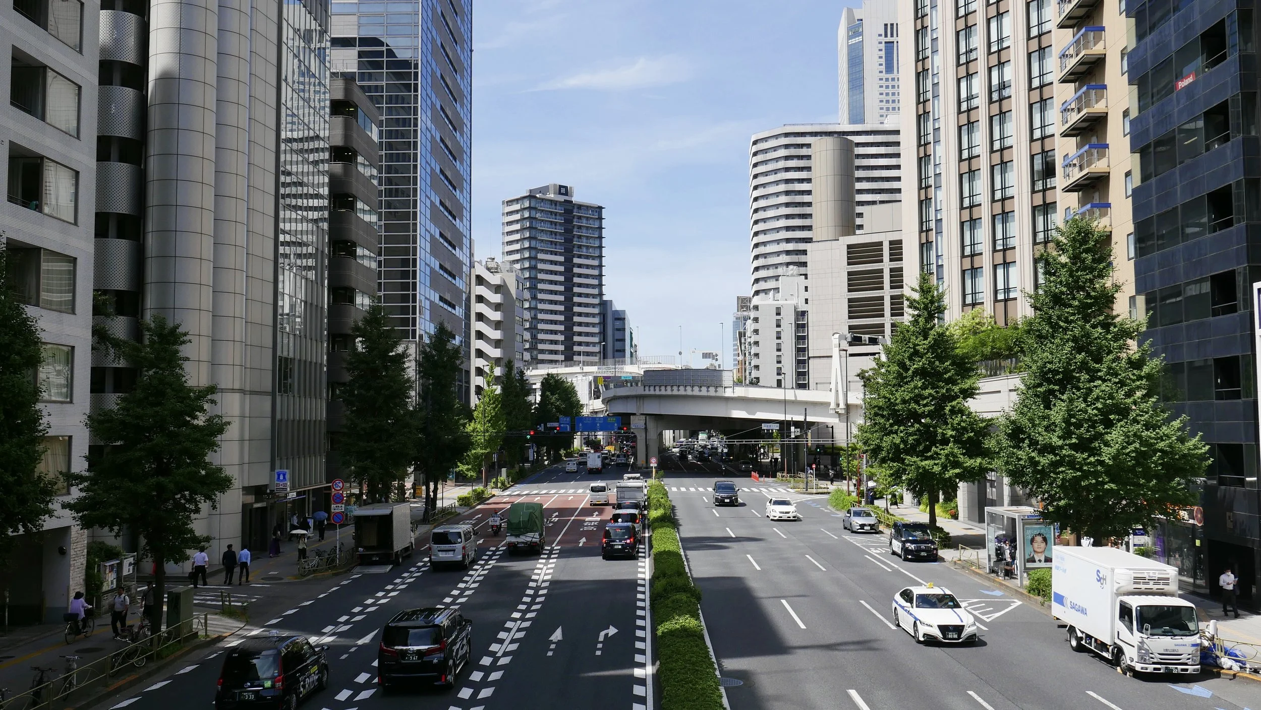 A city street scene with high-rise buildings, cars on the road, and trees lining the sidewalks on a clear day.