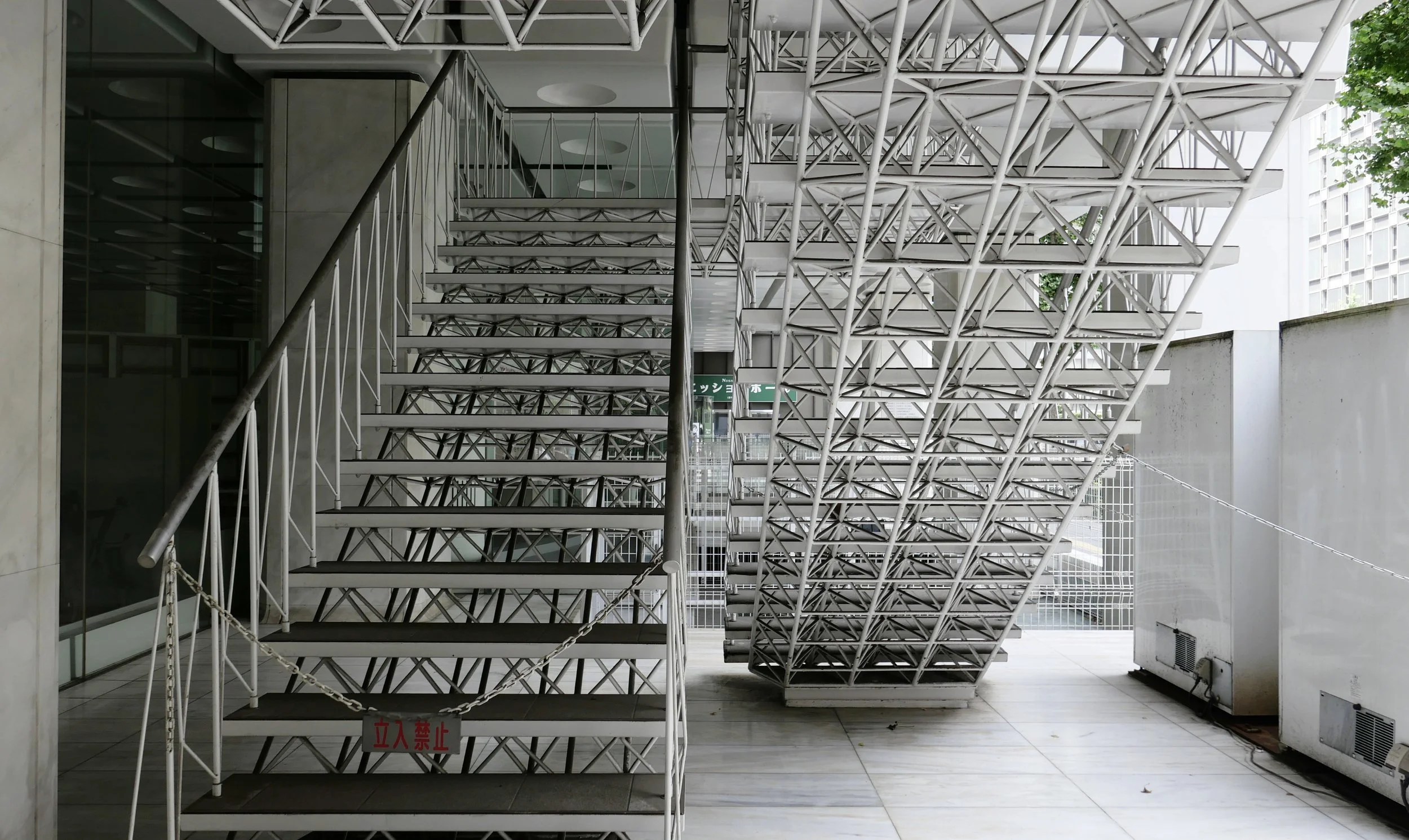 Exterior view of a modern building with white metal stairs and railing, green trees in the background, and Japanese signs visible.