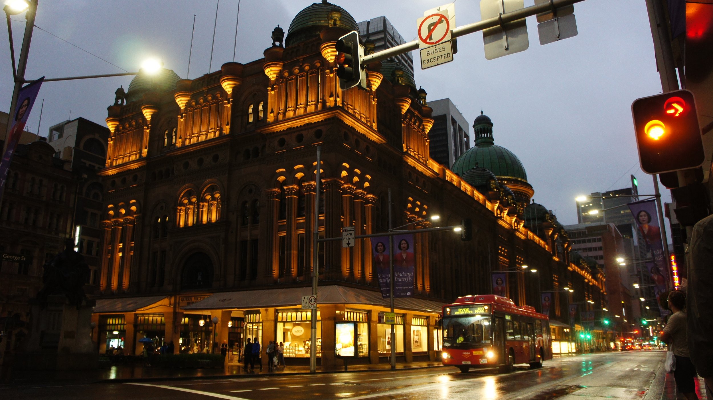 Historic building illuminated at dusk in a busy city street with traffic, pedestrians, and a red bus, on a rainy evening.