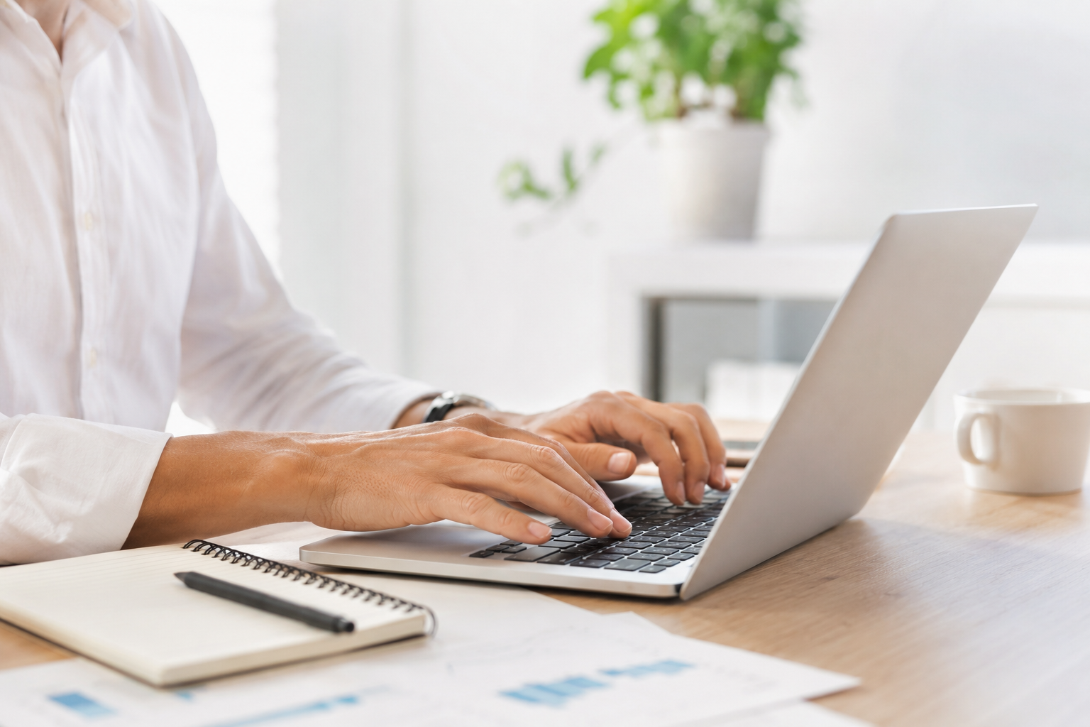 Person working on a laptop at a desk with a notebook, pen, coffee mug, and some papers, in a bright room with a potted plant in the background.