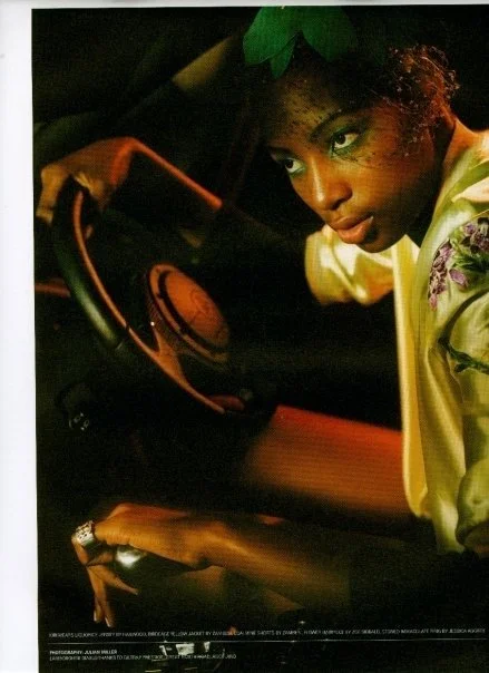 A woman with curly hair, wearing a green hat and a yellow satin jacket, looking over her shoulder inside a car at night.