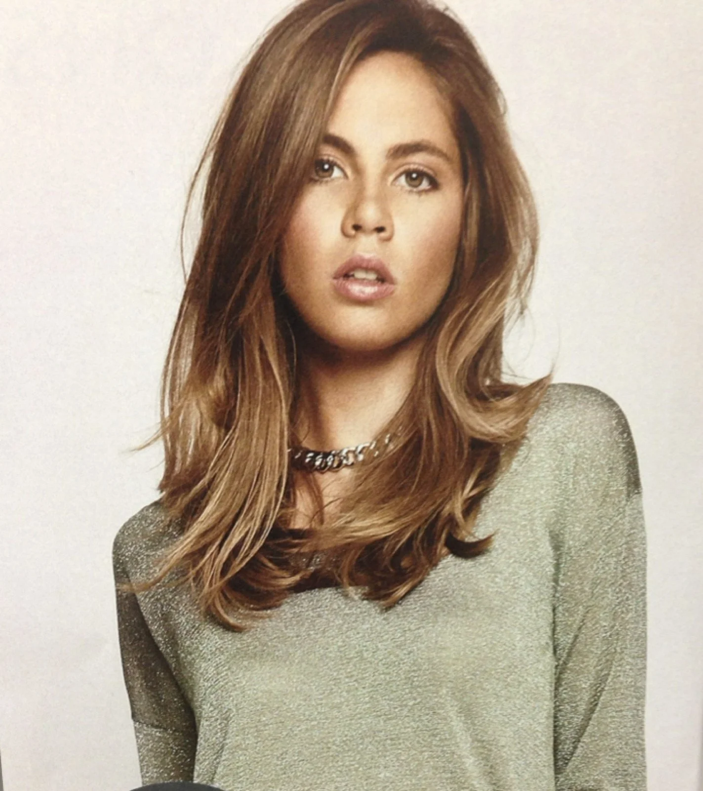 A young woman with long, wavy, light brown hair, wearing a metallic-looking sweater and a silver chain necklace.