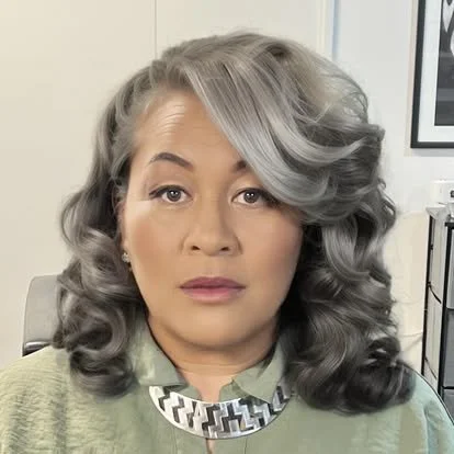 Senior woman with styled gray hair, wearing a green top and a decorative necklace, in an indoor setting.