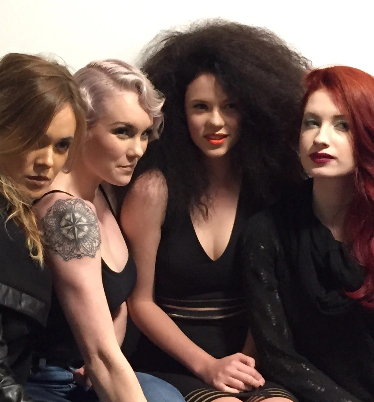 Four women sitting close together, dressed in dark clothing, with varied hairstyles and hair colors, against a plain white background.