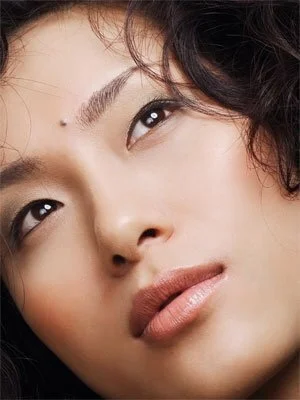Close-up of a woman's face with makeup, dark wavy hair, and a neutral expression.