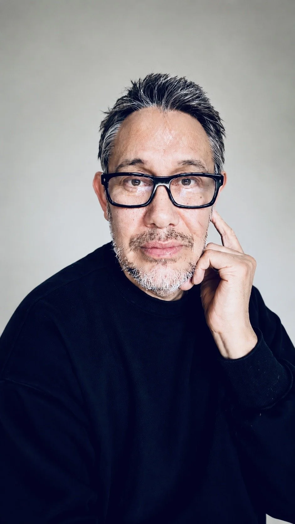 A middle-aged man with glasses, gray hair, and a beard, wearing a black shirt, posing with his hand touching his face against a plain light background.