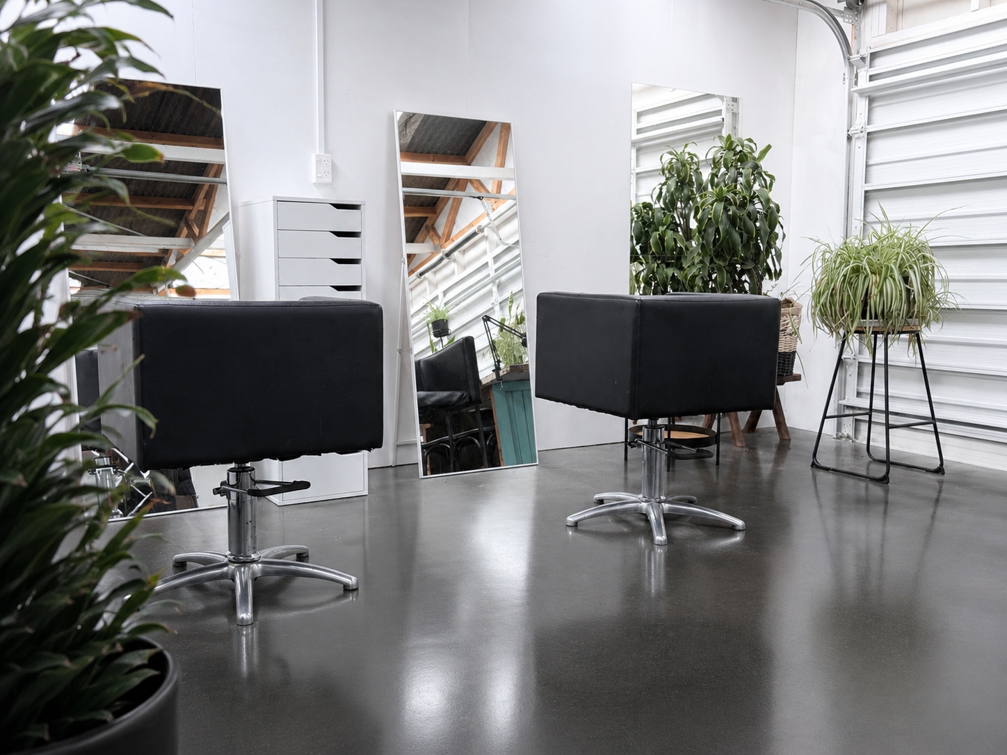 Modern hair salon interior with black styling chairs, large mirrors, and potted plants near a white wall and metal garage door.