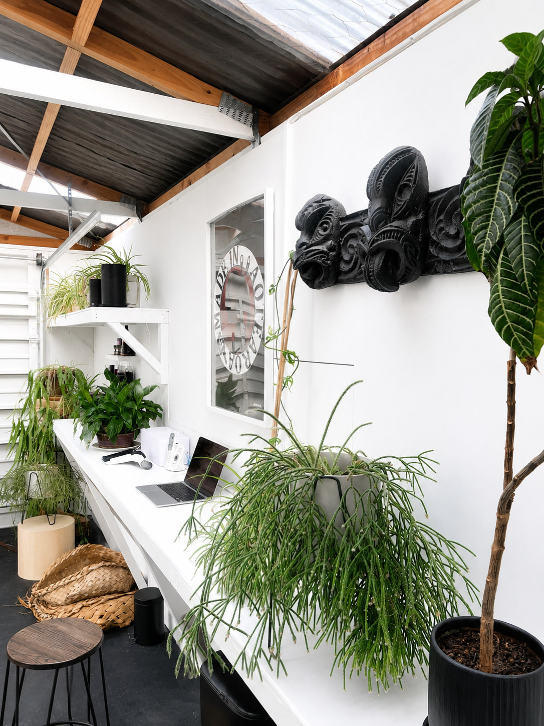 Interior of a modern workspace with white walls, a desk with plants, a laptop, and a mirror, featuring decorative black tribal masks and potted plants on shelves and the floor.