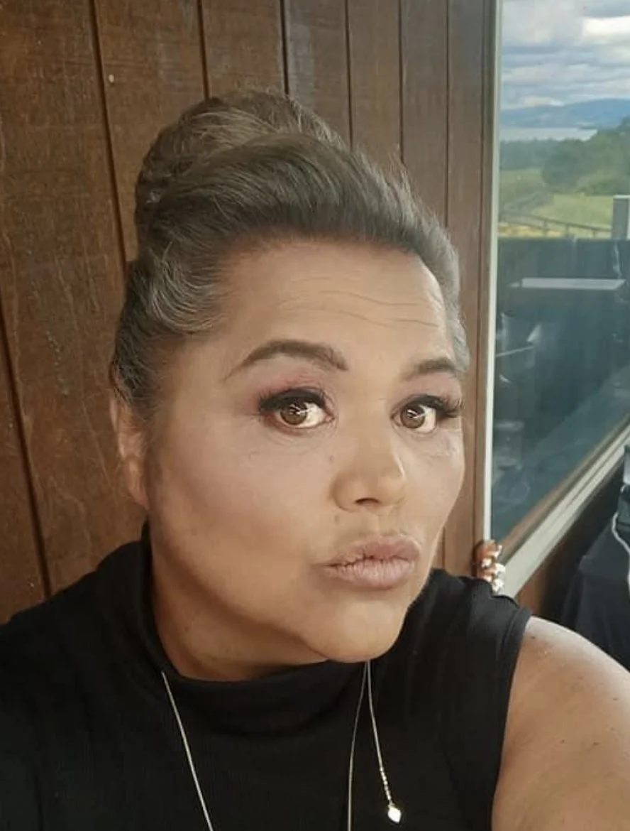 A woman with styled gray hair, wearing makeup and a black sleeveless top, sitting indoors near a window with wood-paneled walls and a scenic landscape view outside.