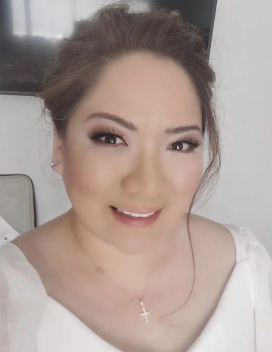 A woman with styled brown hair, makeup, wearing a white top, a cross necklace, and earrings, smiling at the camera indoors.