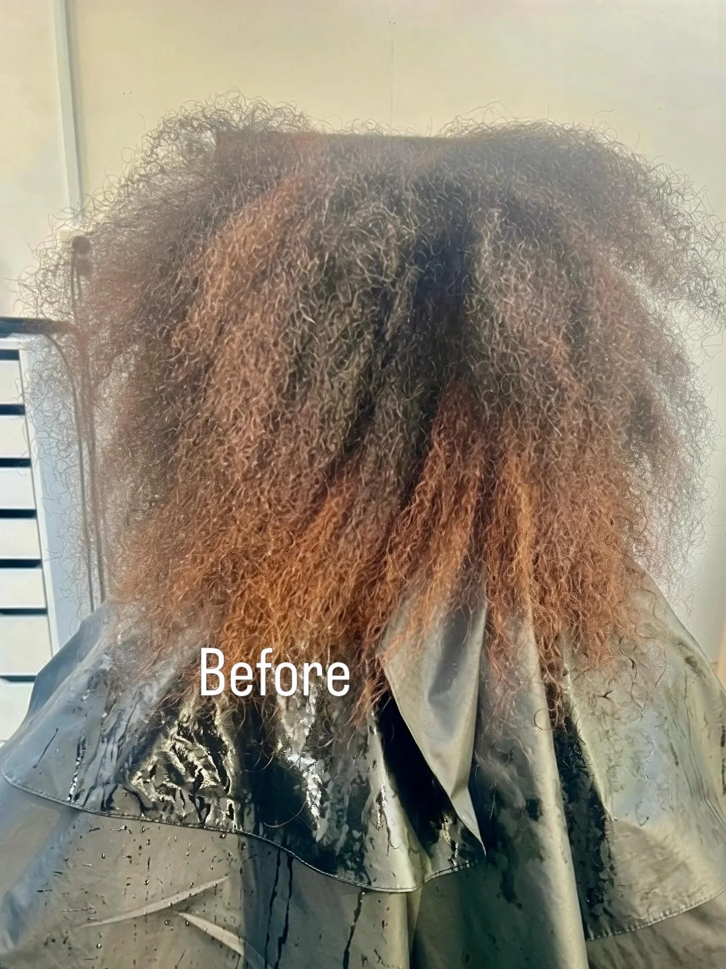 Close-up of a person's hair before a treatment, showing frizzy, curly, and unkempt hair with a mix of dark and light brown shades. The person is wearing a plastic cape typically used in salons.