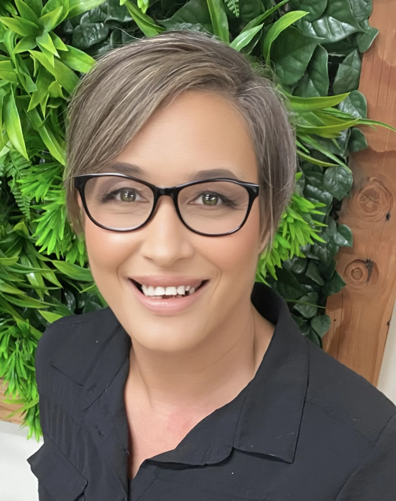 A woman with short blonde hair and glasses smiling, with green leafy plants and a wooden wall in the background.