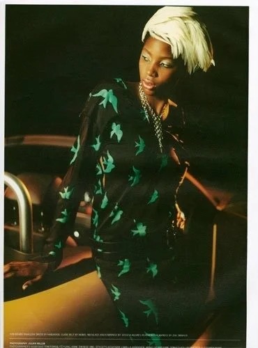 Woman with short platinum blonde hair, wearing a black outfit with green leaf pattern, standing beside a vintage yellow car.