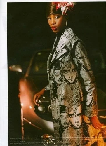 Woman with short hair wearing a patterned coat with illustrated faces, standing in front of a vintage car at night.