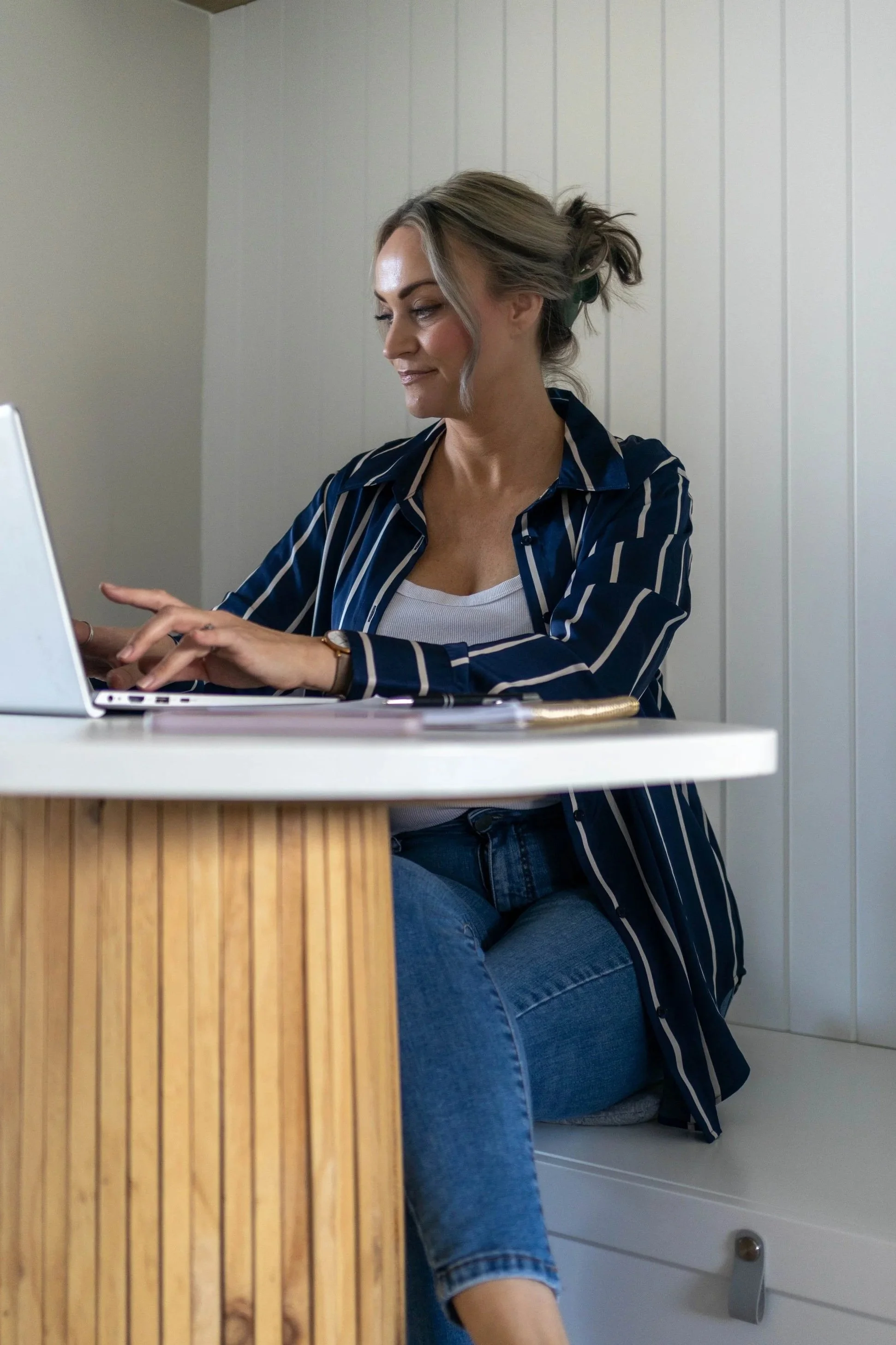 A woman with blonde hair tied back, wearing a striped blue and white shirt and jeans, sitting at a desk working on a laptop, with papers in front of her.