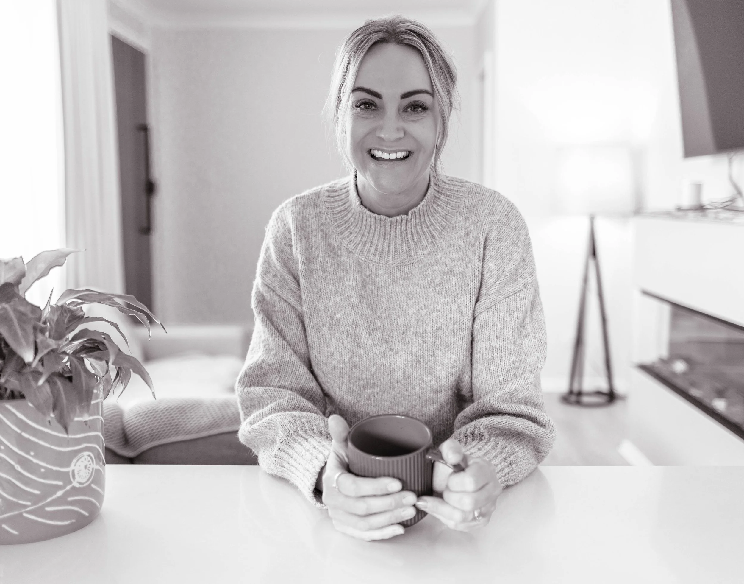 A woman smiling and holding a coffee mug, sitting at a table in a bright room. A large potted plant is in the foreground on the left, and a staircase is visible in the background on the right.