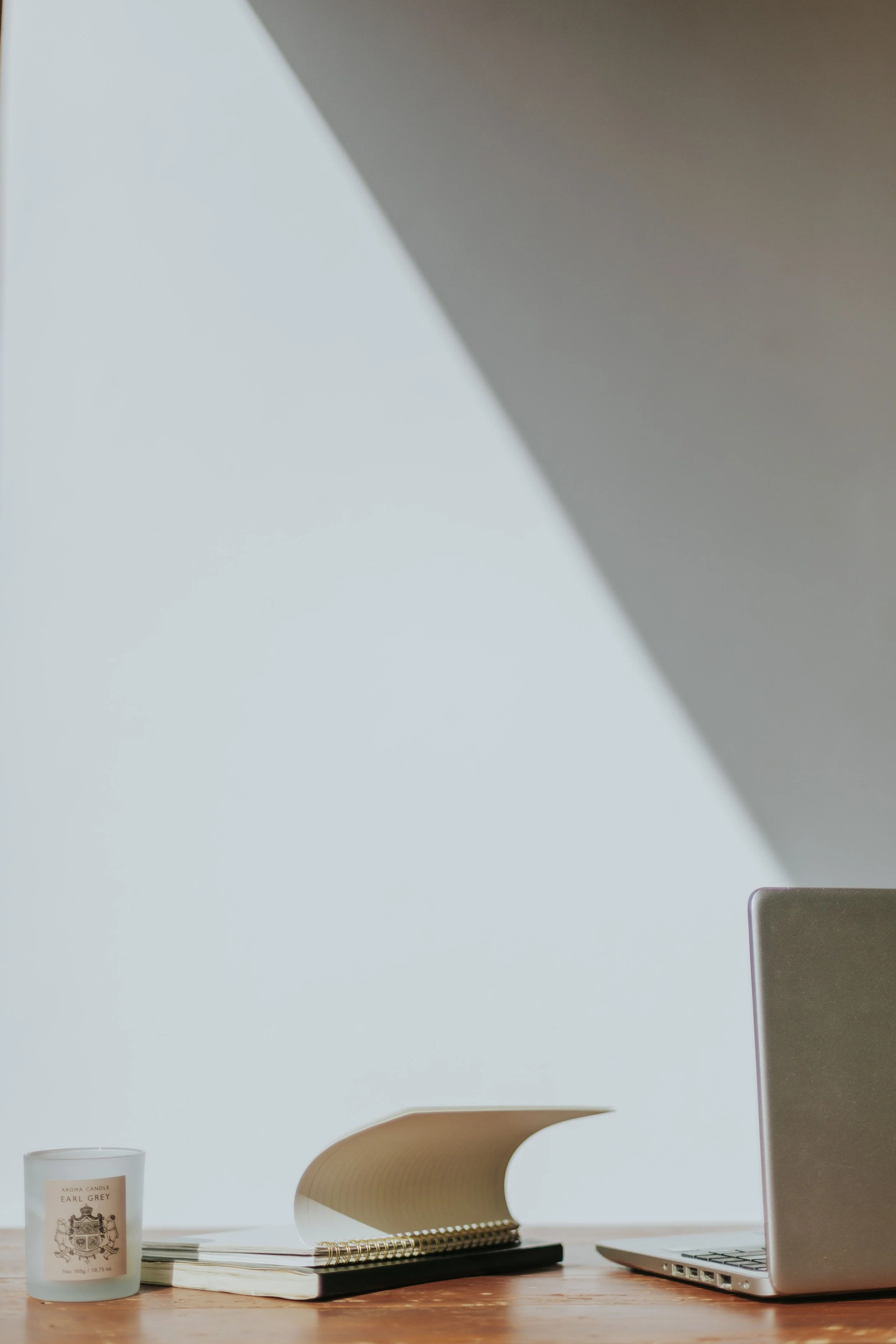 A desk setup with a white candle labeled 'Earl Grey', a closed spiral notebook with a black cover, a tan spiral paper holder, a silver laptop, and a beige ergonomic chair, all against a light wall with a shadow.