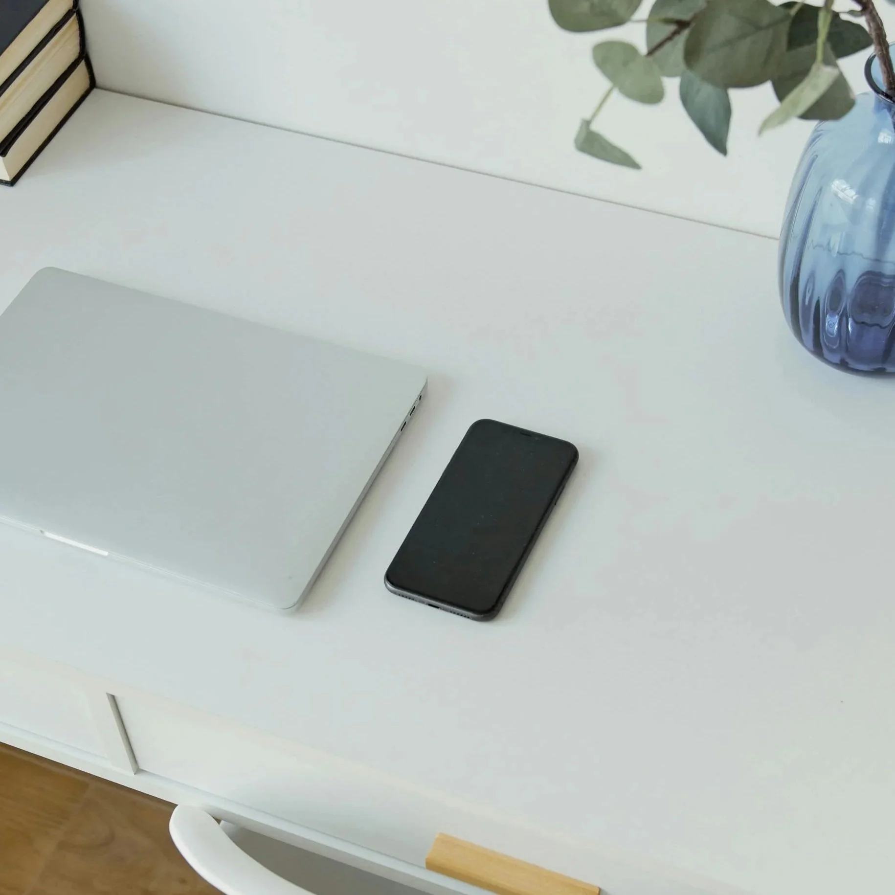 A white desk with a closed silver laptop, a black smartphone, and a blue vase with green leaves in it. A few books are stacked on the left side of the desk.