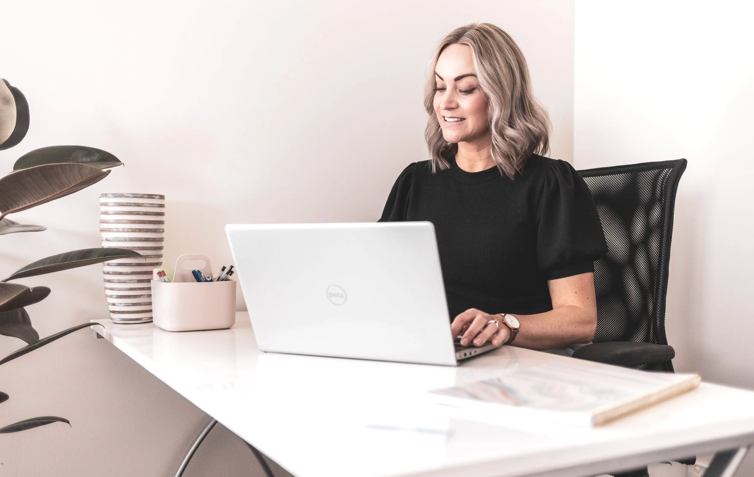 A woman with wavy blonde hair, dressed in black, sitting at a white desk with a Dell laptop, smiling and looking down, in a modern office with a plant, a striped vase, and a container of pens.