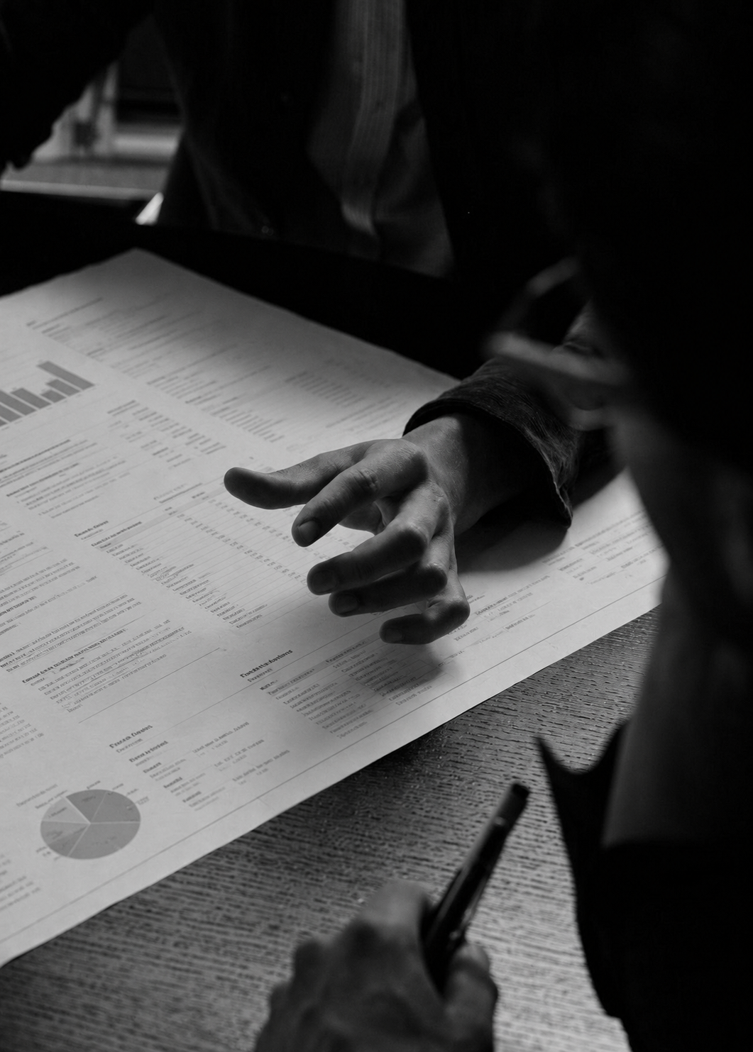 A black and white photo of a person reviewing and taking notes on financial documents with charts and graphs, sitting at a wooden table.