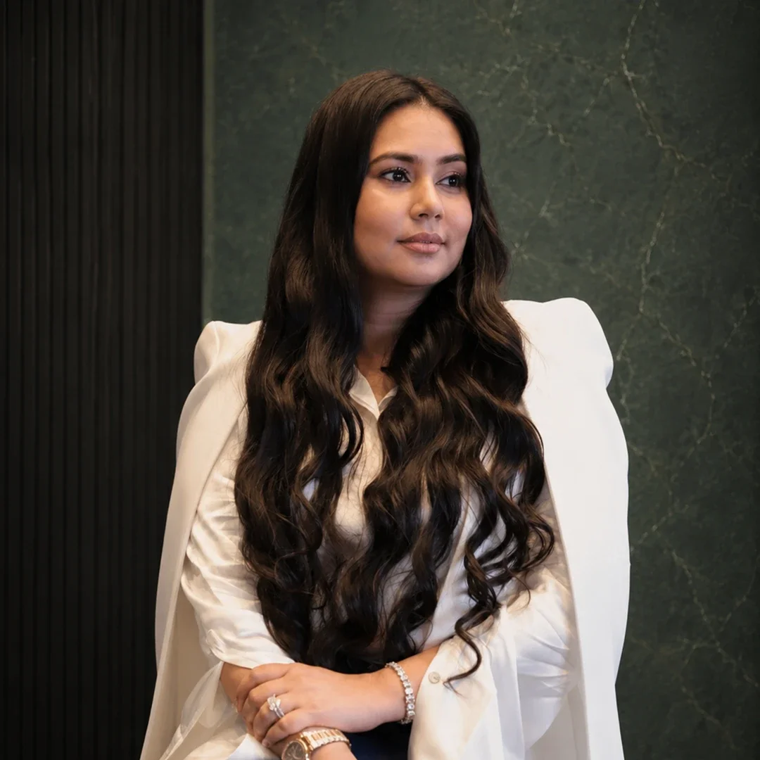 A woman with long dark wavy hair, wearing a white blazer over a cream blouse, sitting with her hands crossed on her lap, against a dark green marble wall.