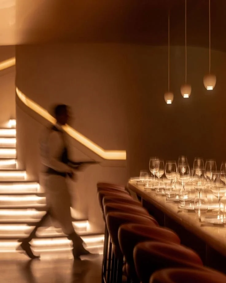 A blurred man walking past a bar with wine glasses and hanging pendant lights, in a dimly lit, modern restaurant or bar interior.