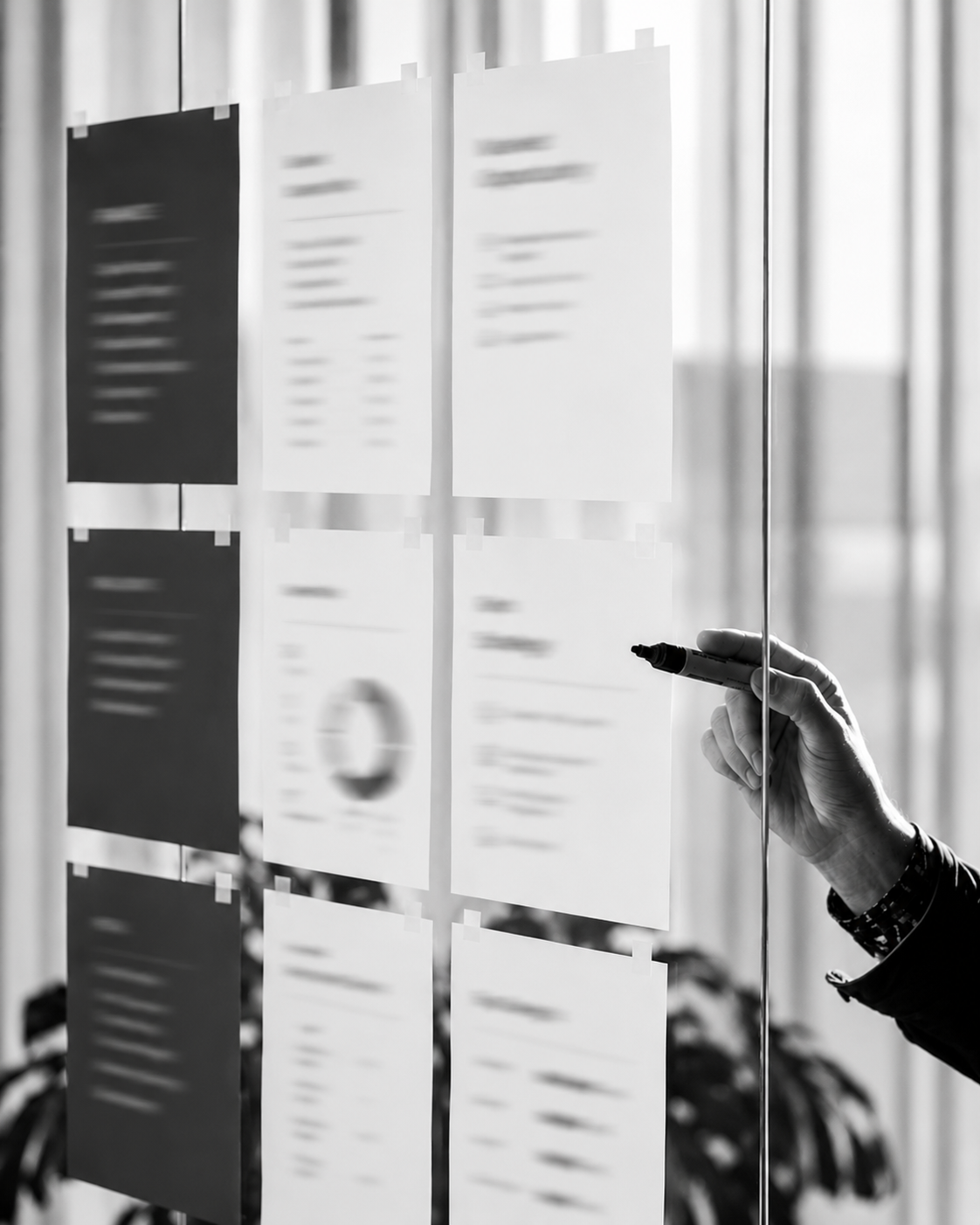 A person writing on a glass wall with papers taped on it, including charts and notes, in a modern office setting.