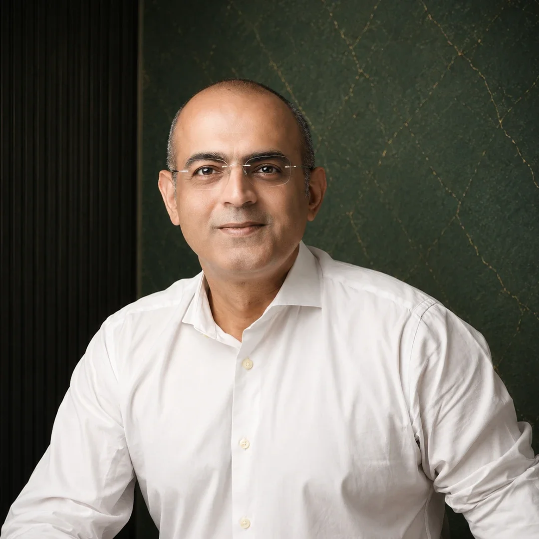 A middle-aged man wearing glasses and a white shirt, sitting in front of a green textured background with gold lines.