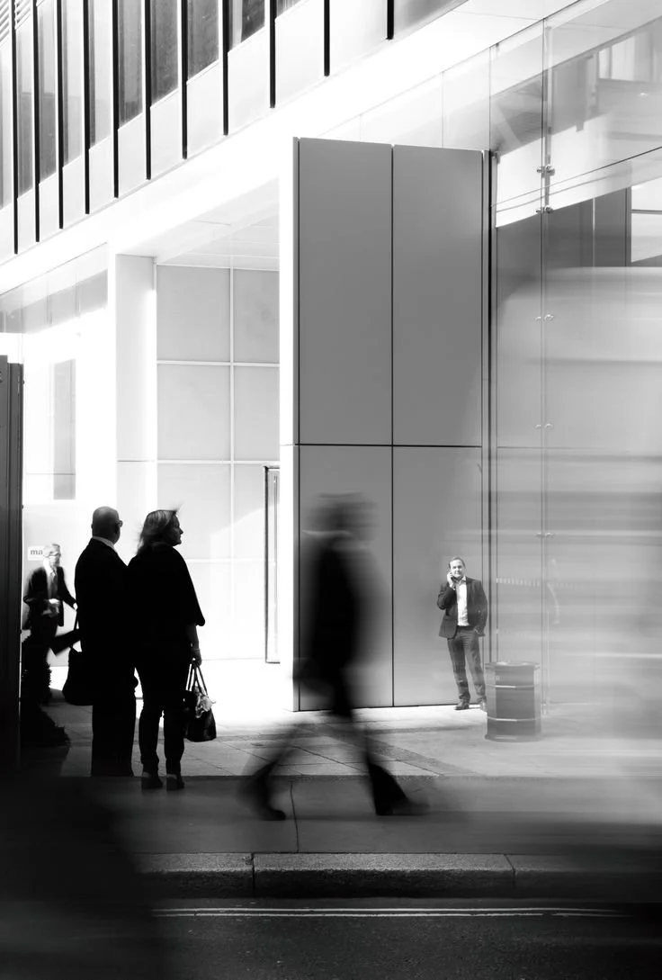 Black and white photo of people inside and outside an office building. Three people standing outside, two of them talking, and one person walking past, blurred. Inside, a man in a suit stands near a reflective glass wall, talking on a cellphone.