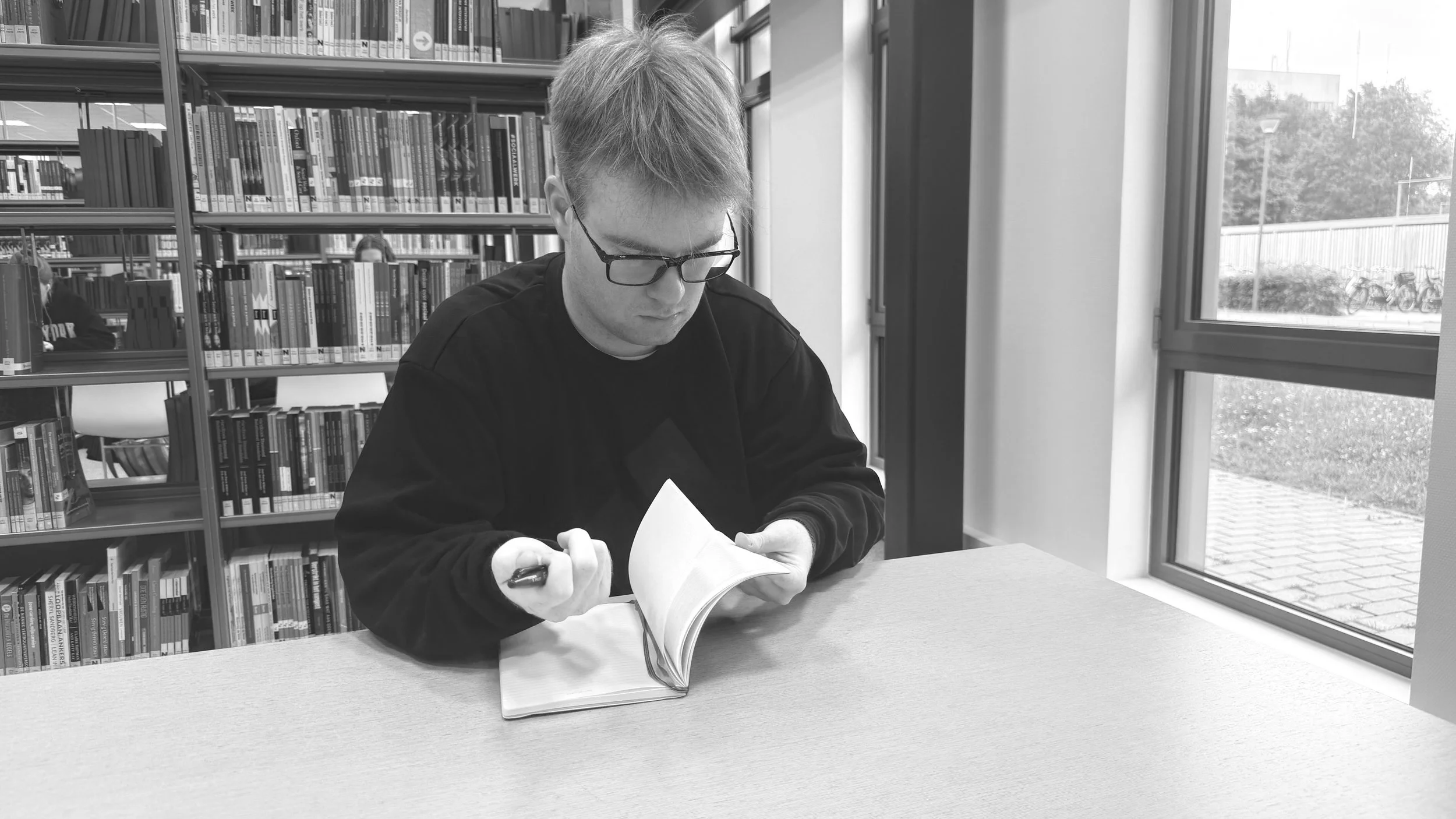 A person with glasses is sitting at a table in a library, reading a book with a pen in hand, near a window overlooking an outside area with trees and bicycles.