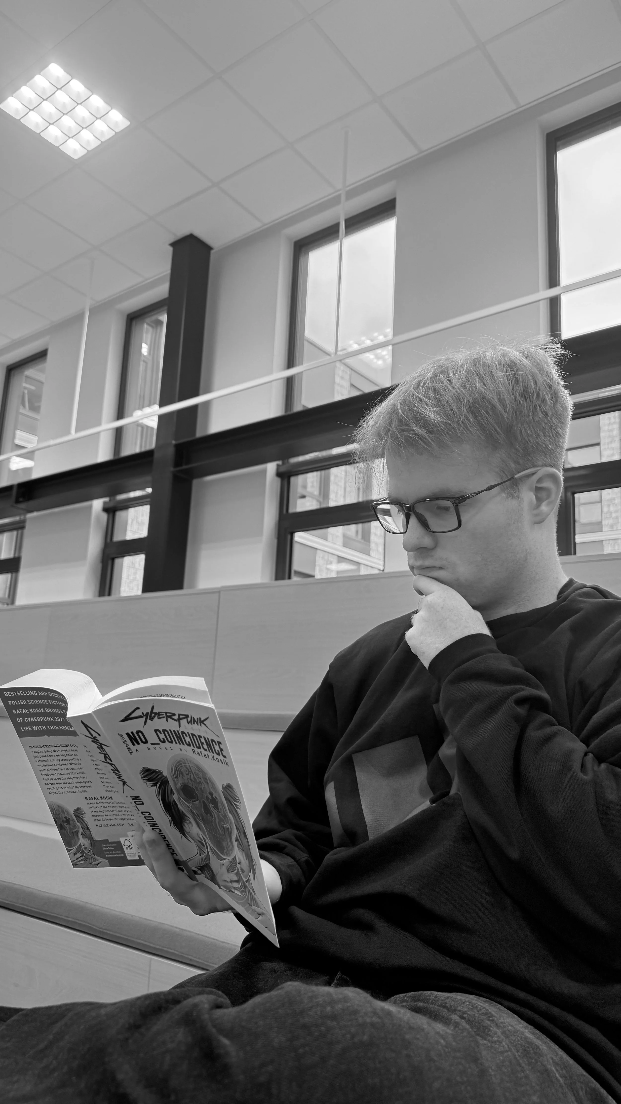 A young man with glasses sits at a table in a modern building, reading a book titled 'Cyberpunk 2077: No Coincidence.' He has light hair and is wearing a dark hoodie. The background features large windows and a high ceiling with square lights.
