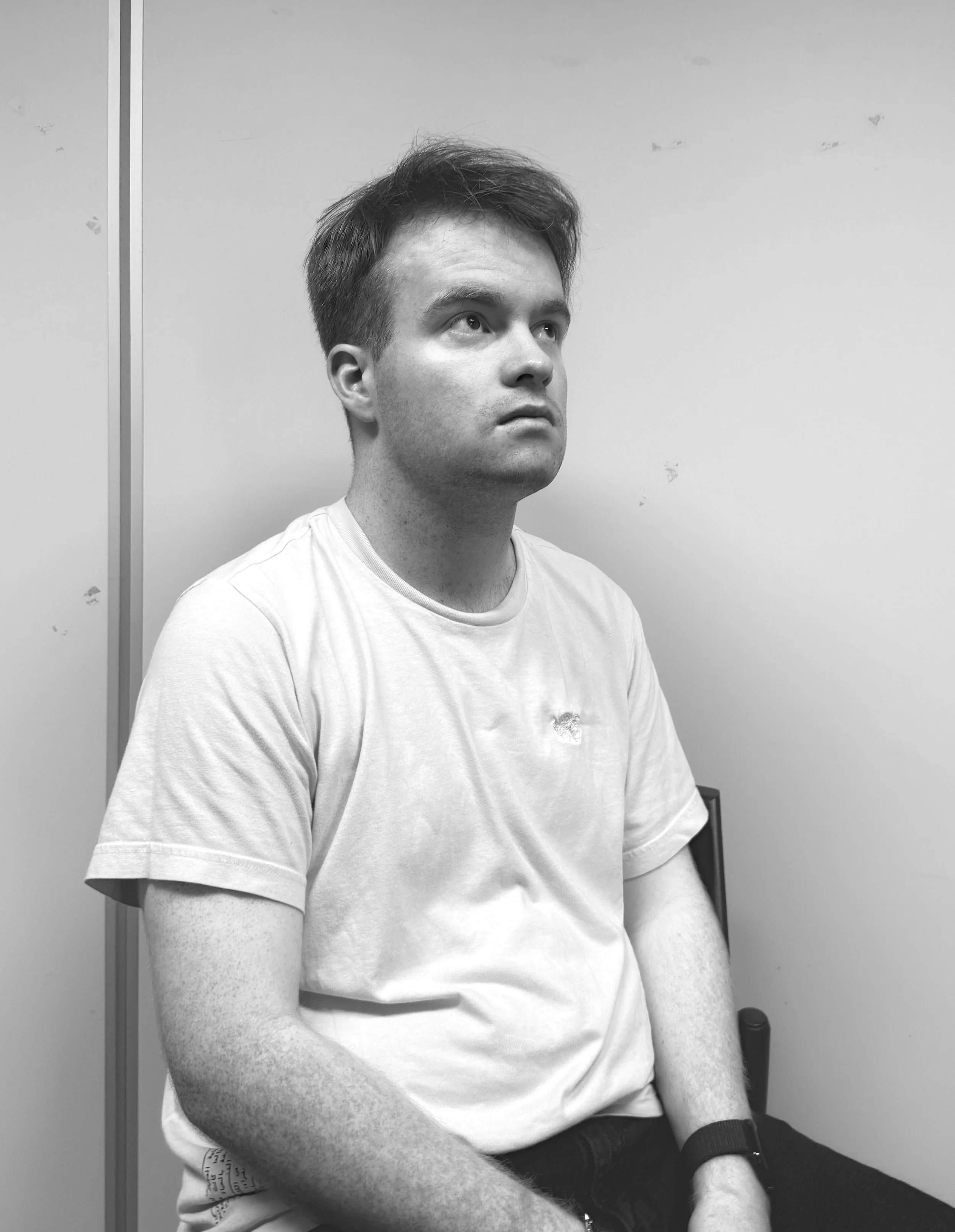 Black and white photo of a young man with short hair, sitting on a chair against a plain wall, looking slightly upward.
