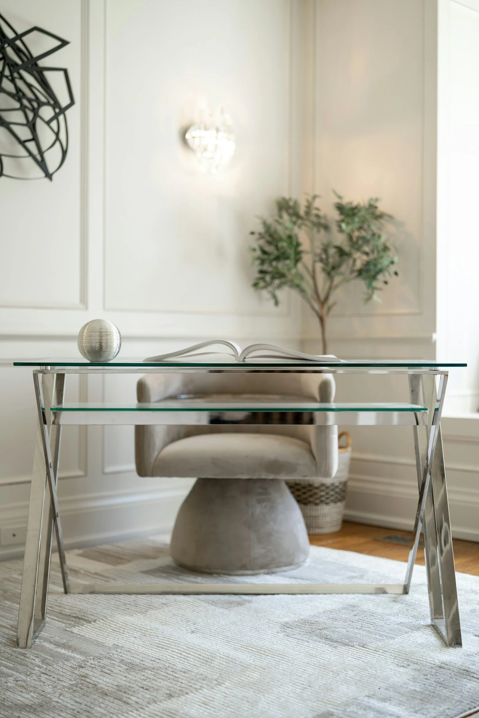 Une table en verre avec un vase en forme de sphère en métal et un livre ouvert. Un fauteuil en velours beige est en dessous de la table, avec un tapis en laine rayé clairs et foncés. En arrière-plan, un mur blanc avec une plante verte et une applique murale