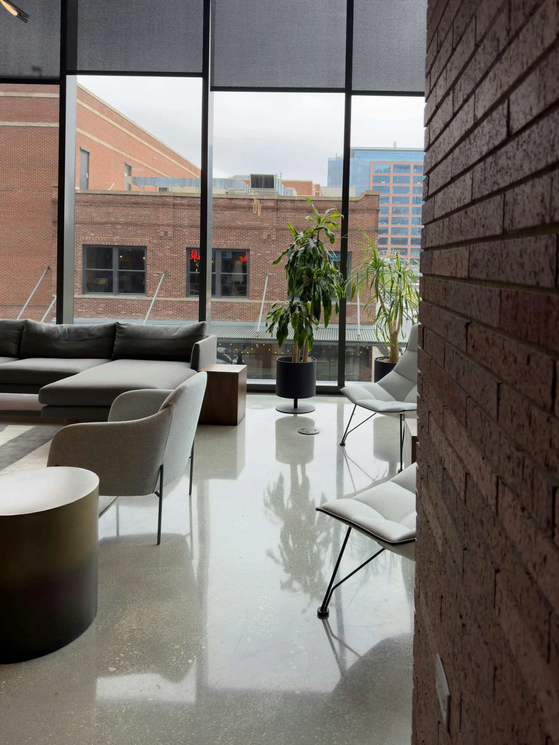 Intérieur moderne avec des fauteuils blancs, une plante verte dans un pot noir, un canapé gris clair, et une grande fenêtre avec vue sur des bâtiments en briques rouges et une structure en verre.